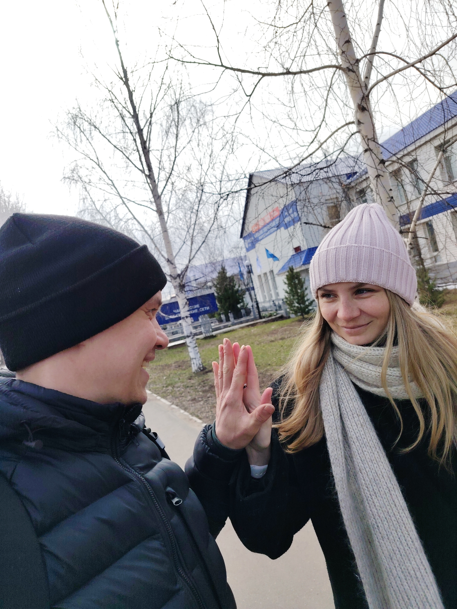 A man and a woman are facing each other outside, with their hands raised in a gesture that could be a high-five or an impending hand clasp. The man is on the left, wearing a black winter hat and a black puffy winter jacket. He is smiling slightly. The woman on the right is wearing a light pink beanie, a gray scarf, and a black coat. She is smiling at the man.
Behind them are trees, some of which are birch trees. In the background is a building with a blue and white facade. A sign on the building contains text in Russian.
Based on the building and the Russian text on the sign, the location is likely in the city of Chaikovsky, Russia.