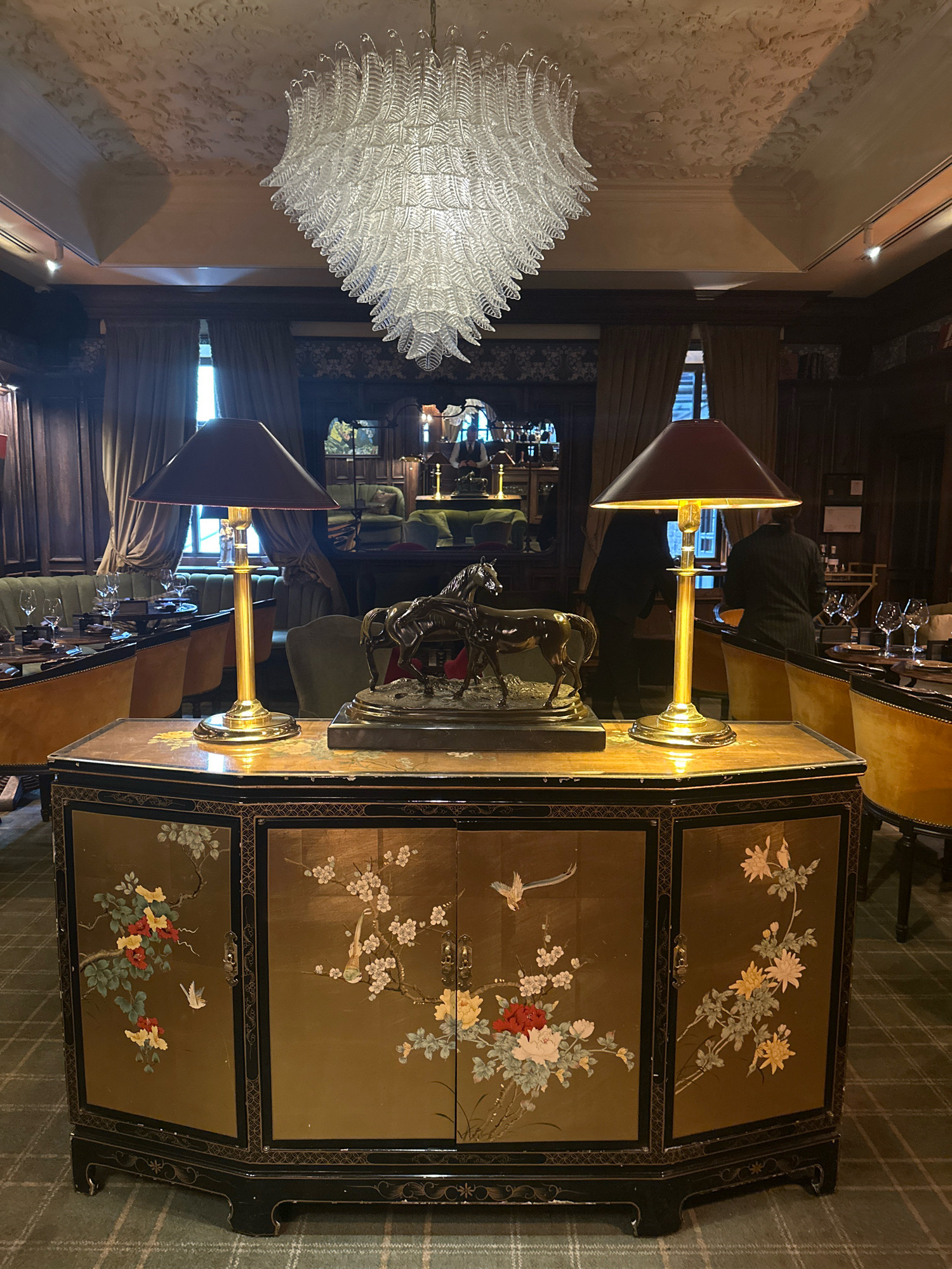 The image depicts an interior scene, likely a restaurant or lounge, featuring an ornate chandelier hanging from a textured ceiling. In the foreground, a dark-colored, lacquered cabinet with intricate floral and bird motifs in gold and color stands on clawed feet. Two brass table lamps with dark conical shades flank a bronze sculpture of horses on top of the cabinet. In the background, plush dining chairs with yellow upholstery surround tables set with glassware, and tall curtains hang by windows. Men in dark suits are visible in the background. The flooring is a green plaid carpet. The scene is lit with warm, ambient light. No specific landmarks or indications of the city of Kyiv, Ukraine are present within the image.