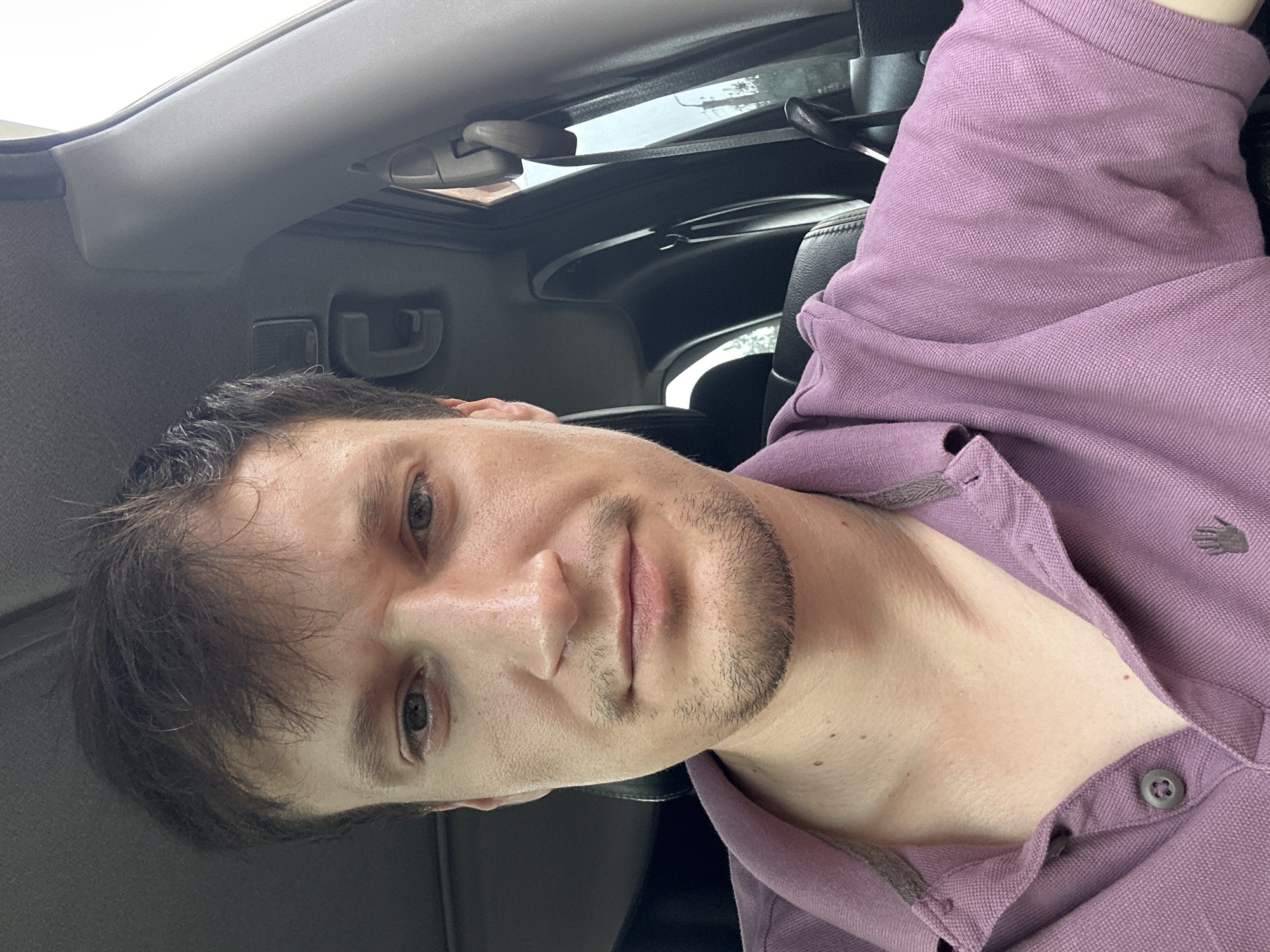 The image shows a close-up, selfie-style shot of a man from the chest up, seated inside a vehicle. The man has light skin, dark brown hair, and is wearing a purple polo shirt. He has a short beard and mustache. The interior of the car is visible behind him, including car seats, a seatbelt, and the car's roof lining. The lighting suggests an outdoor setting, likely daytime. There are no identifiable landmarks to pinpoint the location within Makiv, Ukraine.