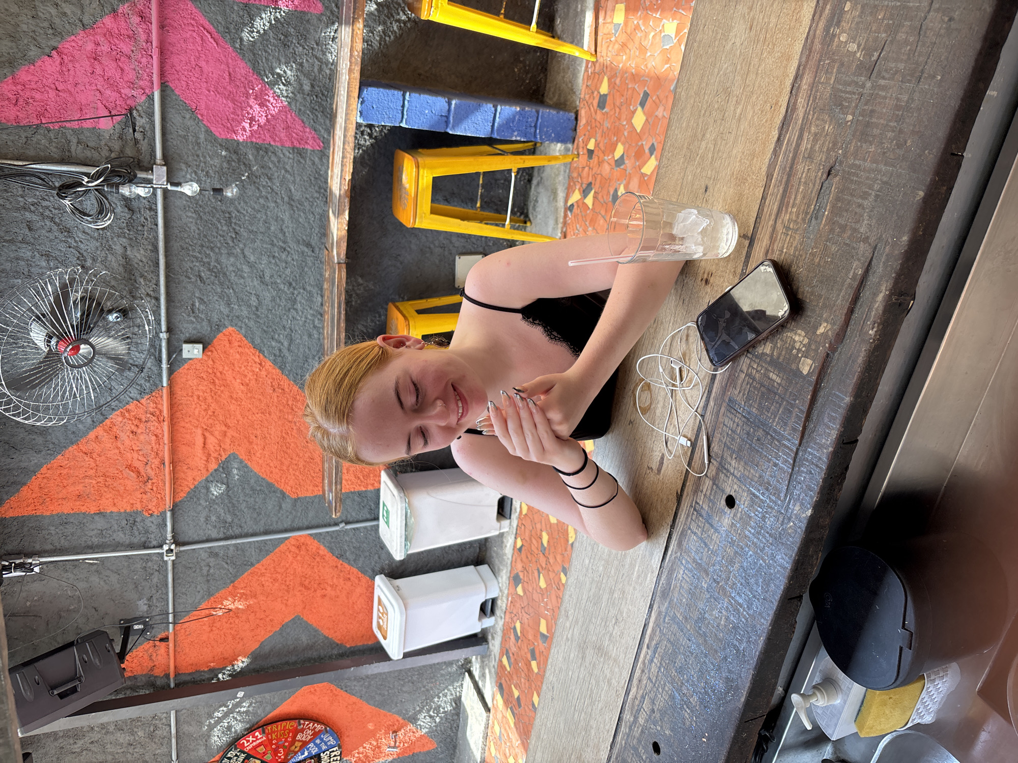A young woman with reddish-blonde hair is seated at a rustic wooden counter, observed from a slightly elevated angle in what appears to be a vibrant, urban cafe or bar in São Paulo, Brazil. She is wearing a black strap top with lace detailing and several black bangles on her left wrist. Her manicured nails are visible as she rests her hands on the counter. She looks down with a gentle, relaxed smile. A clear glass with ice and a white straw, along with a smartphone and tangled white wired earbuds, are placed on the counter near her.
The background features a dynamic setting with dark grey walls adorned with large, bold geometric patterns in orange and pink, resembling triangles or chevrons. Exposed metal pipes and electrical conduits run across these walls, supporting various fixtures like a metal fan, small light bulbs, and a mounted black speaker. Below the counter, the floor is covered in a colorful mosaic of small, irregular tiles in shades of orange, yellow, black, and grey. Two bright yellow stools are visible under the counter, adding a pop of color to the industrial-chic aesthetic. Two white dispensers are mounted on the wall behind the woman. In the bottom right, a section of what looks like a service area includes a black pump dispenser and a sponge. A partially visible circular sign on the lower left wall displays fragmented text, including "2x1", "TRIPLE", "JUMP FREE", "STAMP ON", and "BODY", suggesting promotional offers or event information. The scene is brightly lit, indicating a daytime setting, and exudes a casual, lively atmosphere.