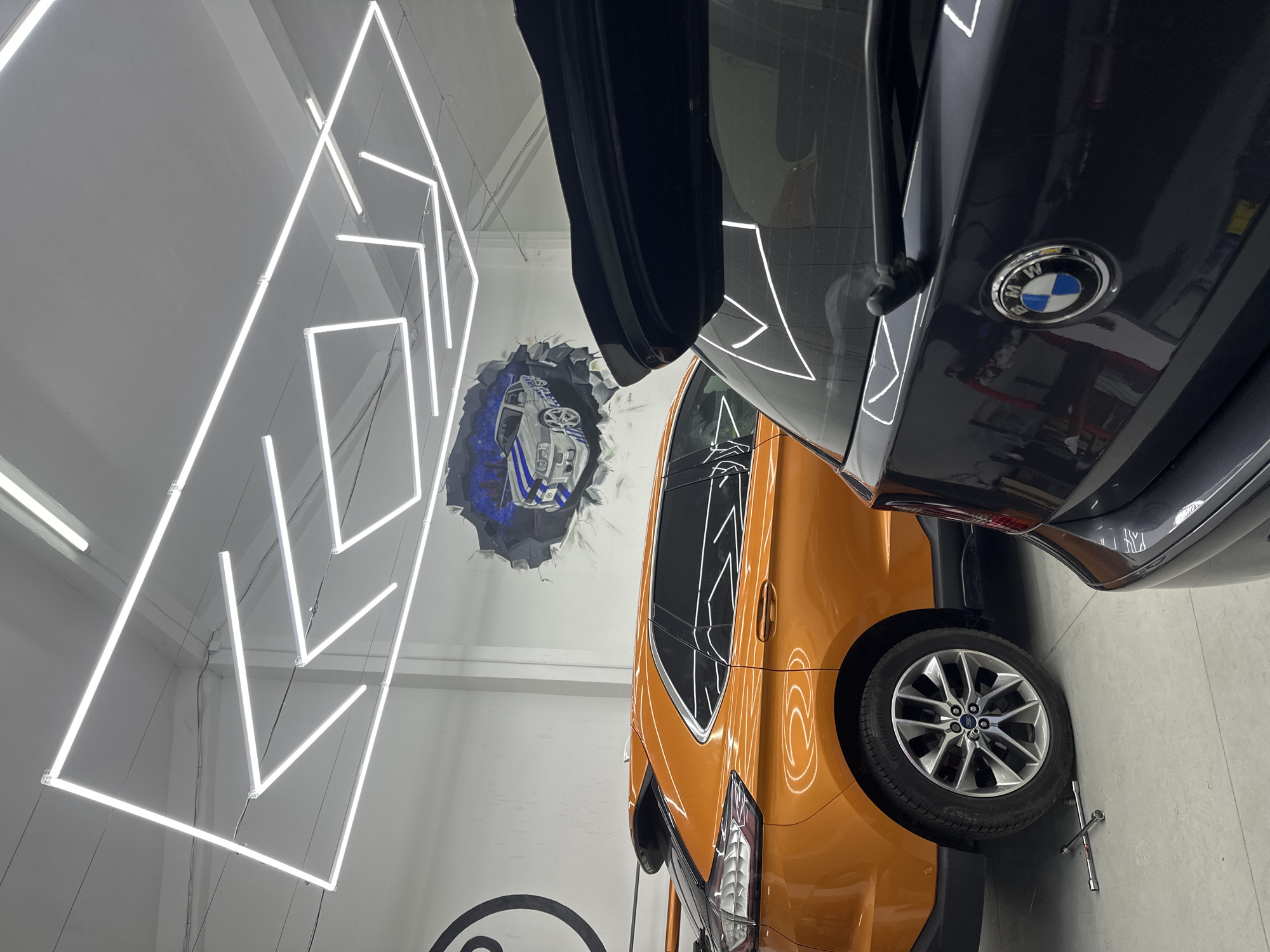 Here is a description of the image:
The image depicts an indoor scene, possibly a car detailing shop or garage, with three cars in the foreground. An orange Ford EcoSport is prominently displayed, parked next to a dark gray BMW with its trunk open. The BMW's badge is visible. A partially visible third car is also present. The cars are on a light gray floor, and a lug wrench is visible near the front wheel of the Ford EcoSport. The background features a white wall decorated with a geometric pattern of illuminated white neon lights. There is also a mural or painting depicting a car bursting through the wall. The environment is brightly lit. There are no people in the scene. The location is Unknown, Unknown.