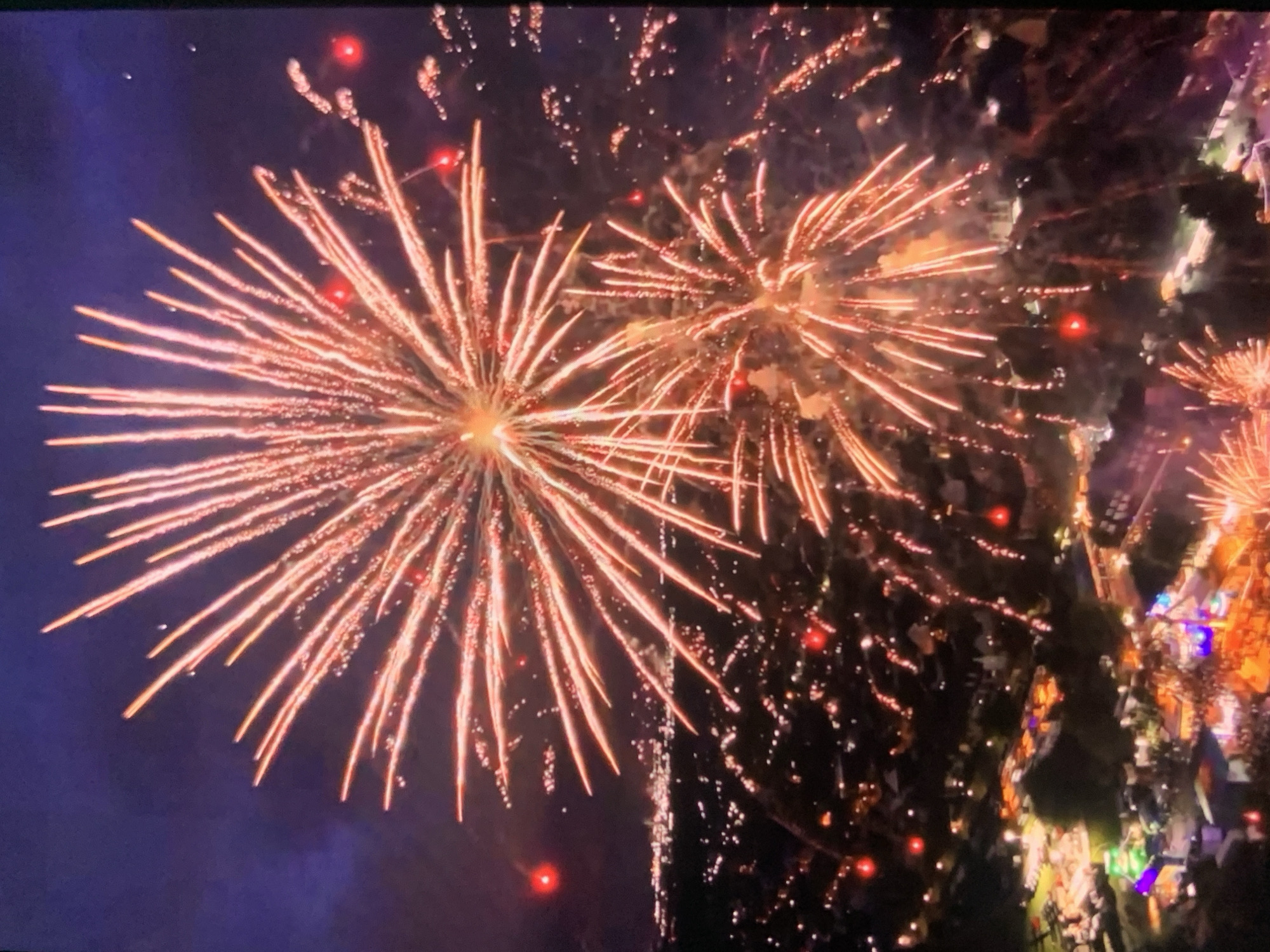 This aerial photograph captures a nighttime fireworks display over a city. The sky is a deep purple-blue, illuminated by the vibrant bursts of fireworks. The main subject of the image is a large, exploding firework in the upper center, with numerous radiant orange and pink streaks extending outwards. Below and to the right of this, a smaller firework is also visible.
In the background, a cityscape is illuminated by scattered lights, suggesting buildings and streetlights. The lights appear as small dots, some reddish and clustered, possibly indicating residential areas or event venues. A hint of a town or suburban landscape is discernible, with darker shapes suggesting trees and structures.
In the lower portion of the image, there's evidence of a festival or event on the ground. Brightly colored lights, including purple and orange, illuminate fairground rides and stalls. A crowd of people can be seen gathered in this area, though they are small and indistinct due to the distance and low light conditions.
The image does not provide any specific visual cues to identify the location as Stavropol, Russia. The description focuses solely on the visible elements of the scene.