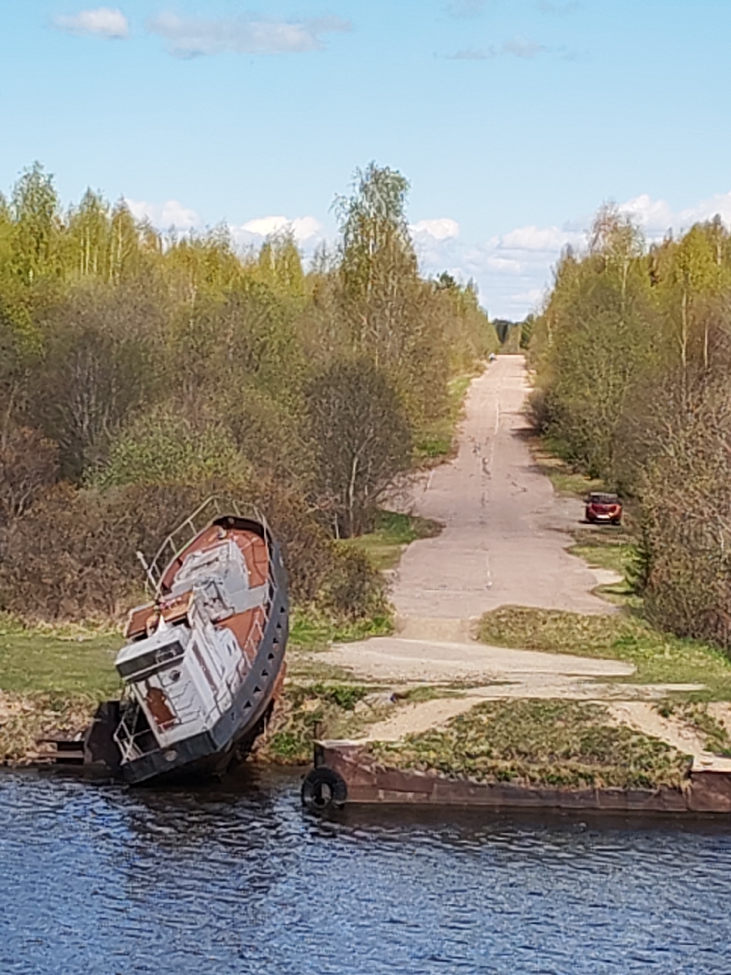 The image shows a waterfront scene near the village of Ivanov Bor, Russia. An old, abandoned boat rests partially submerged at the water's edge. A rusty barge is adjacent to the boat. A deteriorating concrete road stretches into the distance, flanked by dense trees and vegetation. A red car is parked on the right side of the road. The sky is blue with scattered clouds.