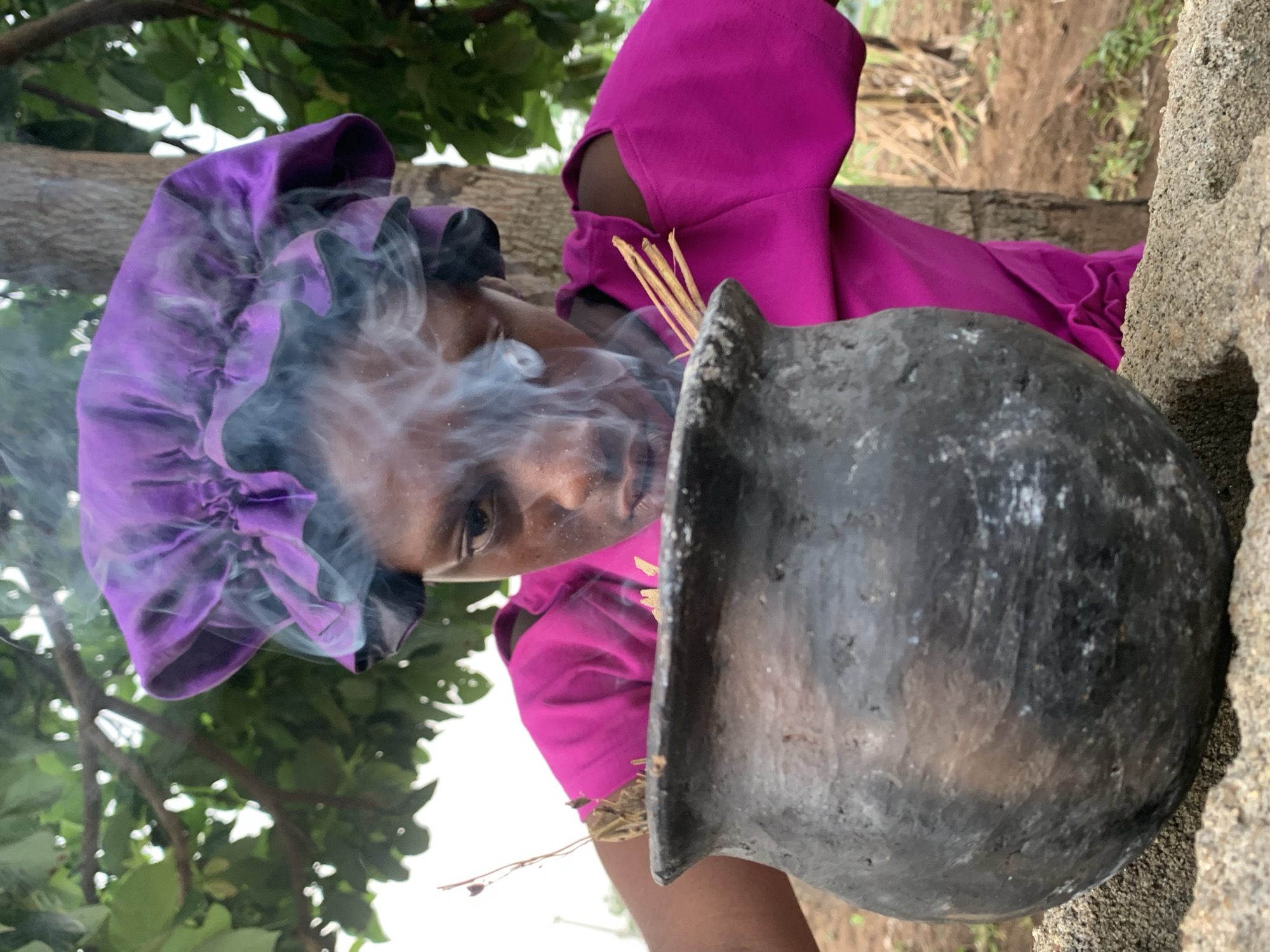 In Agban, Nigeria, a woman wearing a magenta top and purple headscarf is positioned outdoors. She is situated next to a rough concrete structure. In front of her, a black, spherical pot emits smoke; thin sticks are placed near the pot's opening. Trees with green foliage are visible in the background.