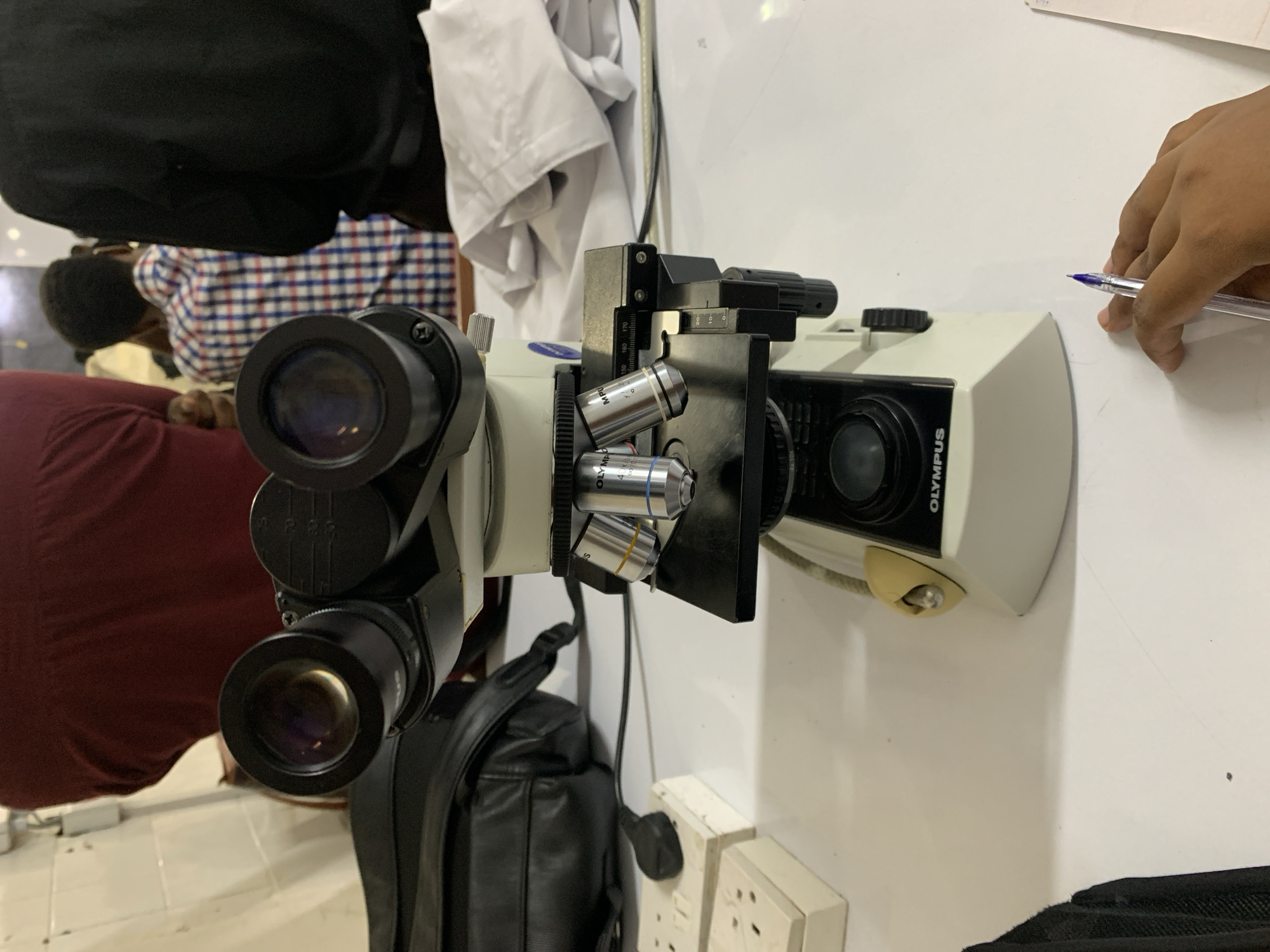 The image depicts an indoor laboratory or classroom setting in Dar es-Salaam, Tanzania. A beige and black Olympus binocular microscope, likely a CX series model, is centrally positioned on a white, reflective table. The microscope features two black eyepieces, a mechanical stage, and a revolving nosepiece displaying four objective lenses, one clearly marked "OLYMPUS 40x/0.65" and another "MPU." A black power cord extends from the microscope's base. To the left of the microscope, a black bag, possibly a shoulder bag or backpack, and two off-white electrical multi-sockets are visible on the table. In the bottom right foreground, a hand, belonging to an individual, holds a clear blue ballpoint pen over a white sheet of paper. The blurred background reveals parts of three individuals: one in a maroon garment, another in a blue and white checkered shirt, and a third in a black garment. A crumpled white lab coat or similar fabric lies on the table behind the microscope, and a tiled wall with white square tiles forms the furthest background. The scene suggests a practical laboratory or educational session in progress.