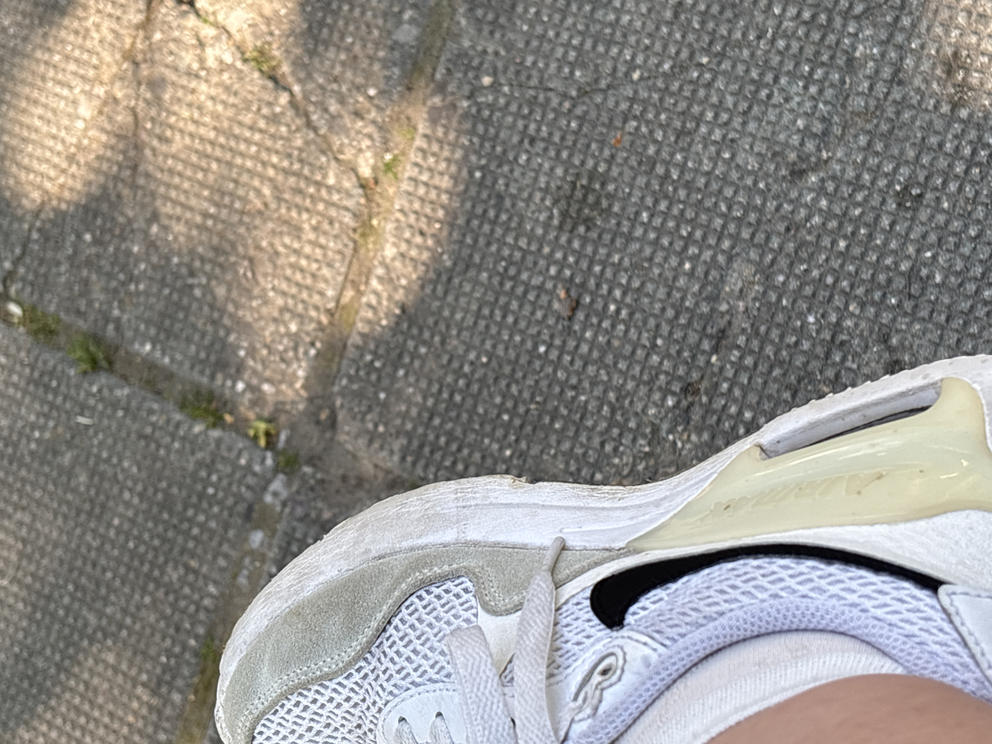 An overhead shot captures a person's foot adorned in a white sneaker, resting on a textured concrete pavement under bright daylight in Gotse Delchev, Bulgaria.
The primary subject is the right foot, partially visible, clad in a white athletic shoe. The shoe features a mesh upper, white laces, and a slightly worn-looking light gray suede-like panel around the toe and midfoot. A small black Nike swoosh logo is discernible on the lateral side of the shoe. The heel area incorporates a prominent translucent, yellowish-tinted air unit, typical of certain sneaker designs. Faint, stylized text, likely "AIR" or a branding element, is subtly molded into this unit. The white sole and upper show signs of everyday wear, with minor scuffs and a slightly dingy appearance in areas. A white sock is visible around the ankle, and a sliver of skin peeks out from the top of the shoe.
The setting is an outdoor paved surface, likely a sidewalk or path, composed of concrete pavers with a distinct grid or square pattern. Sections of the pavement are bathed in bright sunlight, while others are cast in shadow, suggesting an overhead obstruction like a tree or building. Small cracks in the concrete and sporadic patches of green grass or weeds add to the outdoor environment. The strong contrast between light and shadow indicates a sunny day, likely during morning or afternoon hours. No specific activity beyond standing or being present is depicted. There are no discernible emotions or clear textual elements beyond the faint branding on the shoe.