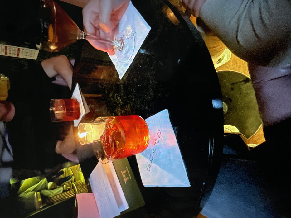 A dark, round table hosts drinks, napkins, and menus. A wine glass is held over a white napkin. Two glasses filled with reddish liquid rest on other napkins, one next to a lit candle. A dark green menu is visible, along with a stack of paper menus and what looks like a receipt book or wallet. Numbers are scrawled on one of the napkins. People surround the table.