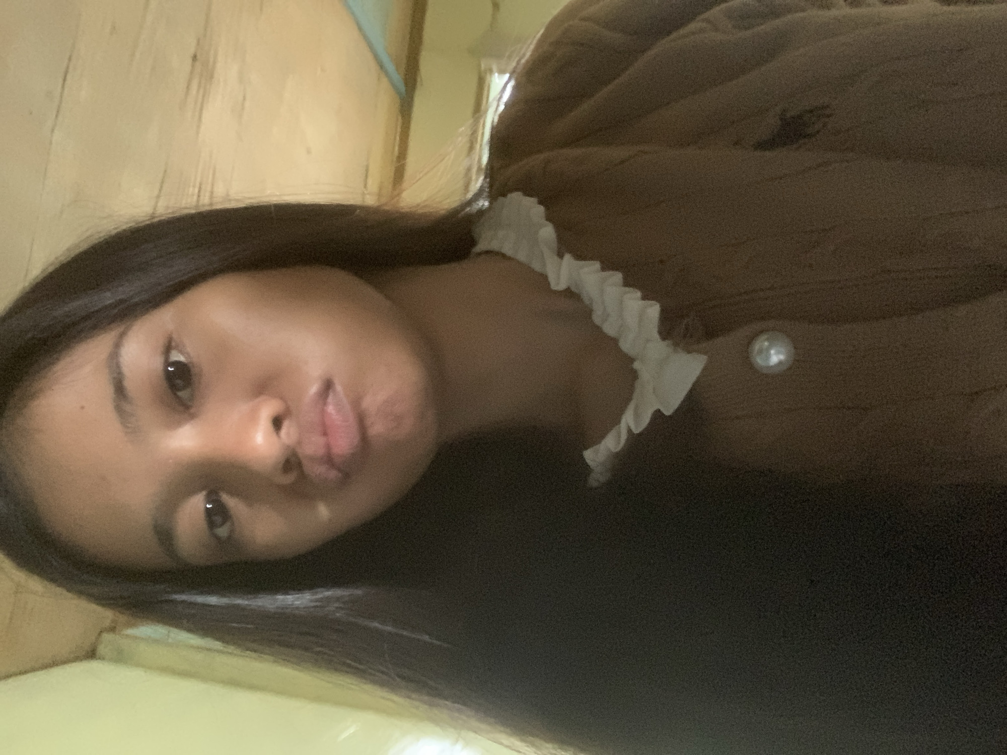 A young woman with dark hair is looking at the camera with her lips pursed. She is wearing a brown sweater with a white ruffled collar and a pearl button. The background appears to be indoors with a wooden ceiling. There are no visible landmarks or contextual clues to determine the specific location within Sungai Meriam, Indonesia.