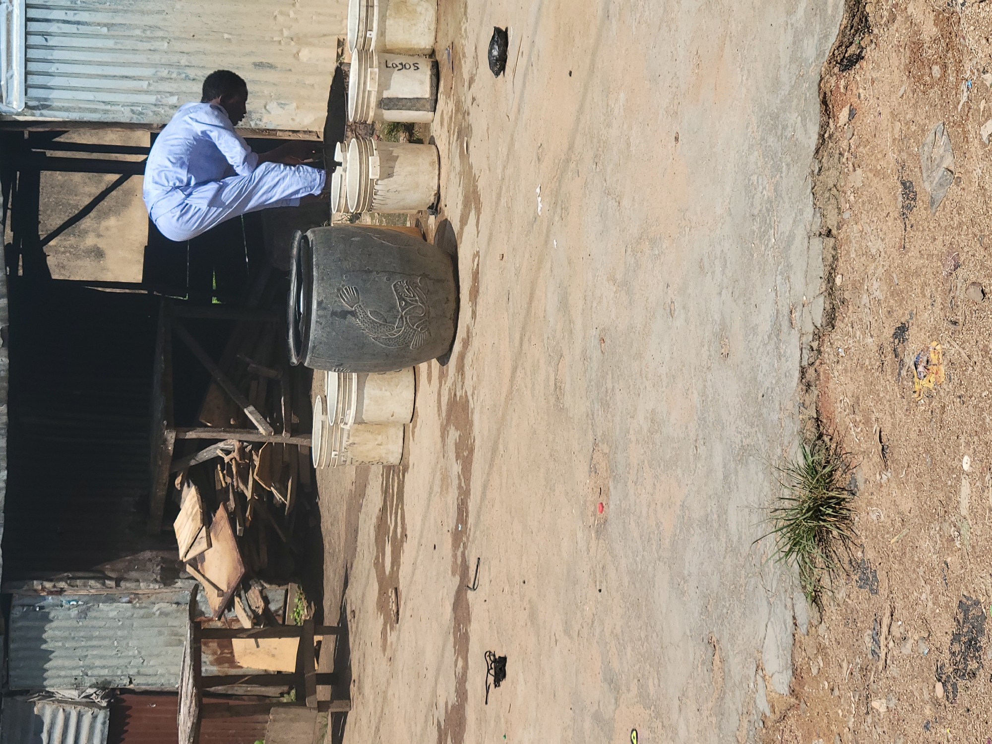 In the city of Jos, Nigeria, a man dressed in a light blue garment is bending over in front of a large, dark gray cylindrical container. The container has a carved image of a fish on its side. Behind the man and the container are several white buckets, some with visible labels, and a pile of wooden planks. The scene appears to be outdoors, with a weathered, corrugated metal structure in the background and a concrete ground surface that shows signs of wear and dirt. There are also scattered debris and a patch of dry grass in the foreground.