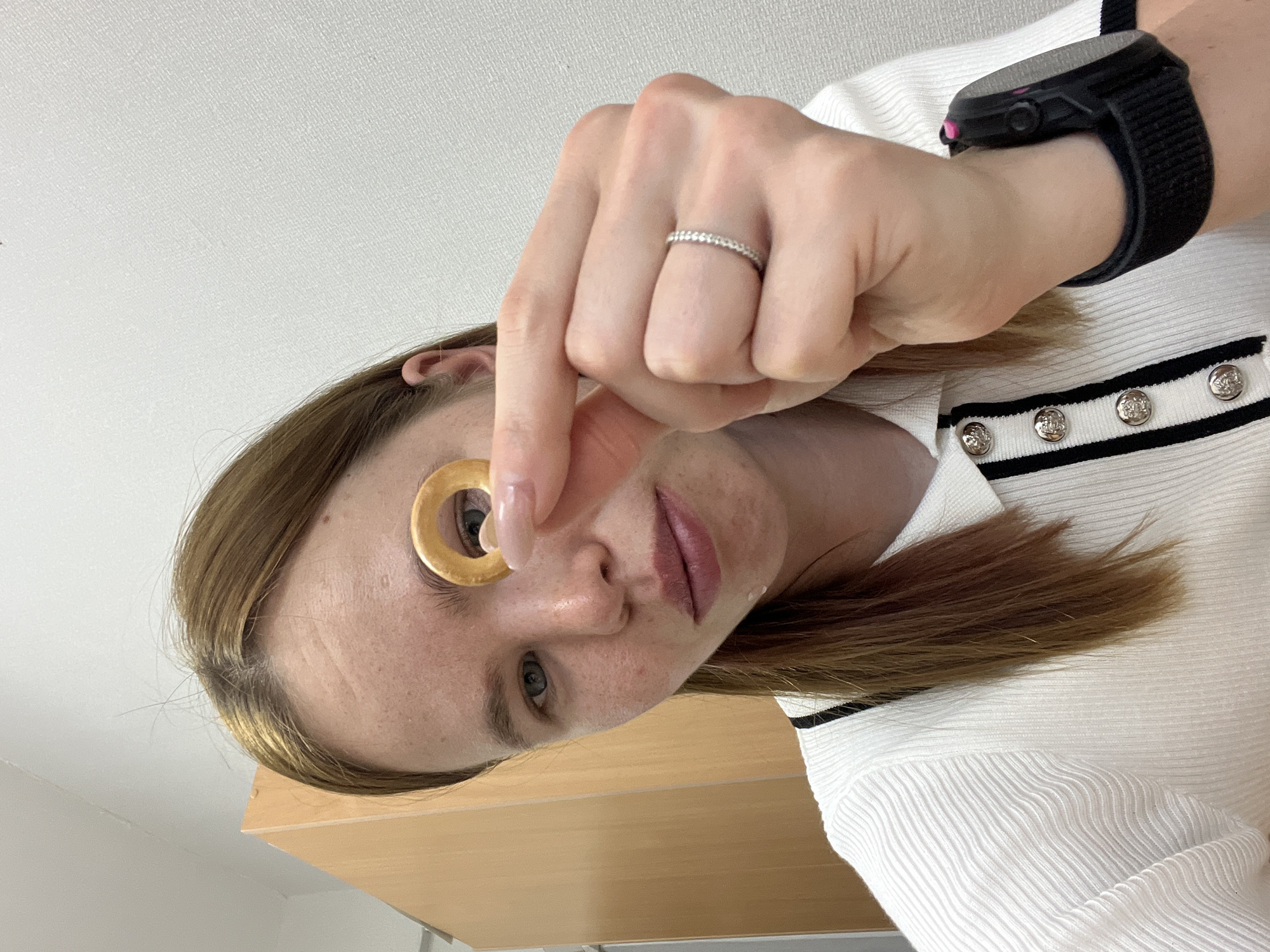 A woman with light brown hair is holding a golden ring up to her left eye. She has fair skin and is wearing a white, ribbed shirt with black stripes and silver buttons. A black smart watch is on her left wrist, and a diamond ring is on her left ring finger. The background is a white ceiling and a light-colored wooden desk or shelf. The location is unknown and could not be determined based on the image.