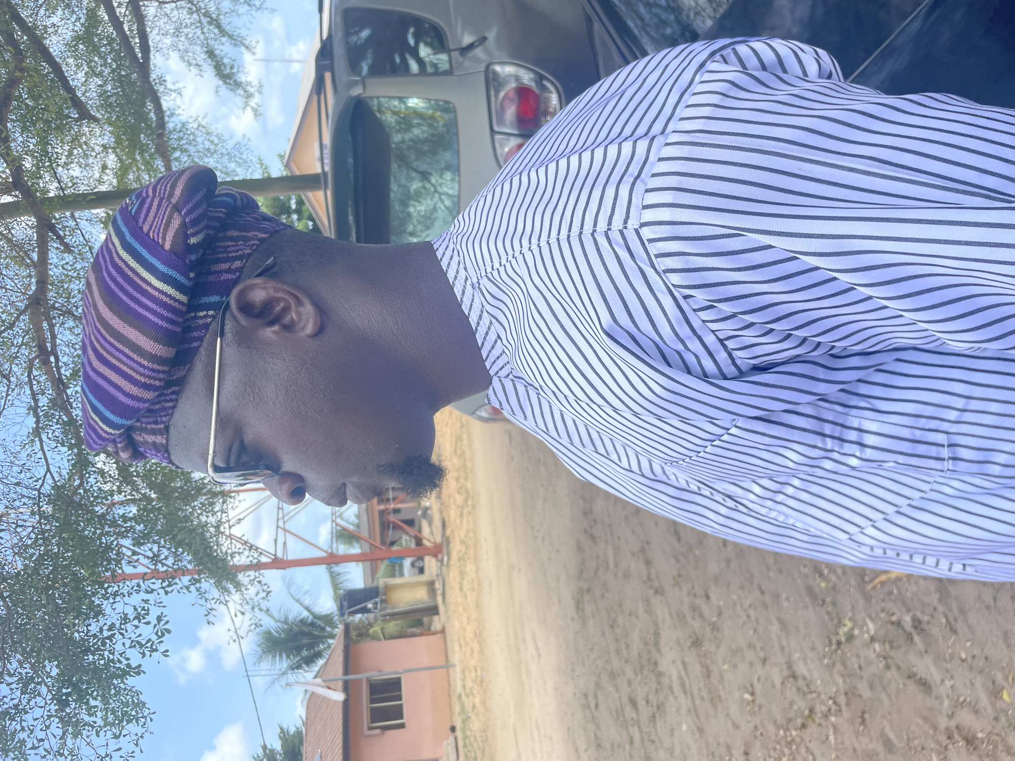 A side view shows a man in Badagry, Nigeria, wearing a striped head covering and a striped shirt. He is wearing glasses with thin gold frames. Behind him is a light-colored SUV with its taillight visible. Trees with sparse foliage are visible on the left side of the frame. A pink building is in the background to the left with an orange metal structure visible above it. The ground is sandy.