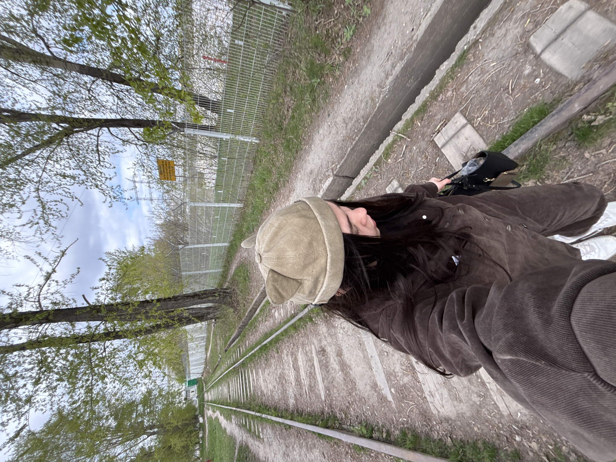 A woman wearing a brown corduroy jacket and a beige hat with ear flaps stands on a narrow-gauge railway line in Perm, Russia. The railway is flanked by green grass and trees on one side and a chain-link fence on the other. A concrete path runs alongside the fence. The woman is holding a black handbag and wearing white sneakers. The sky is overcast.