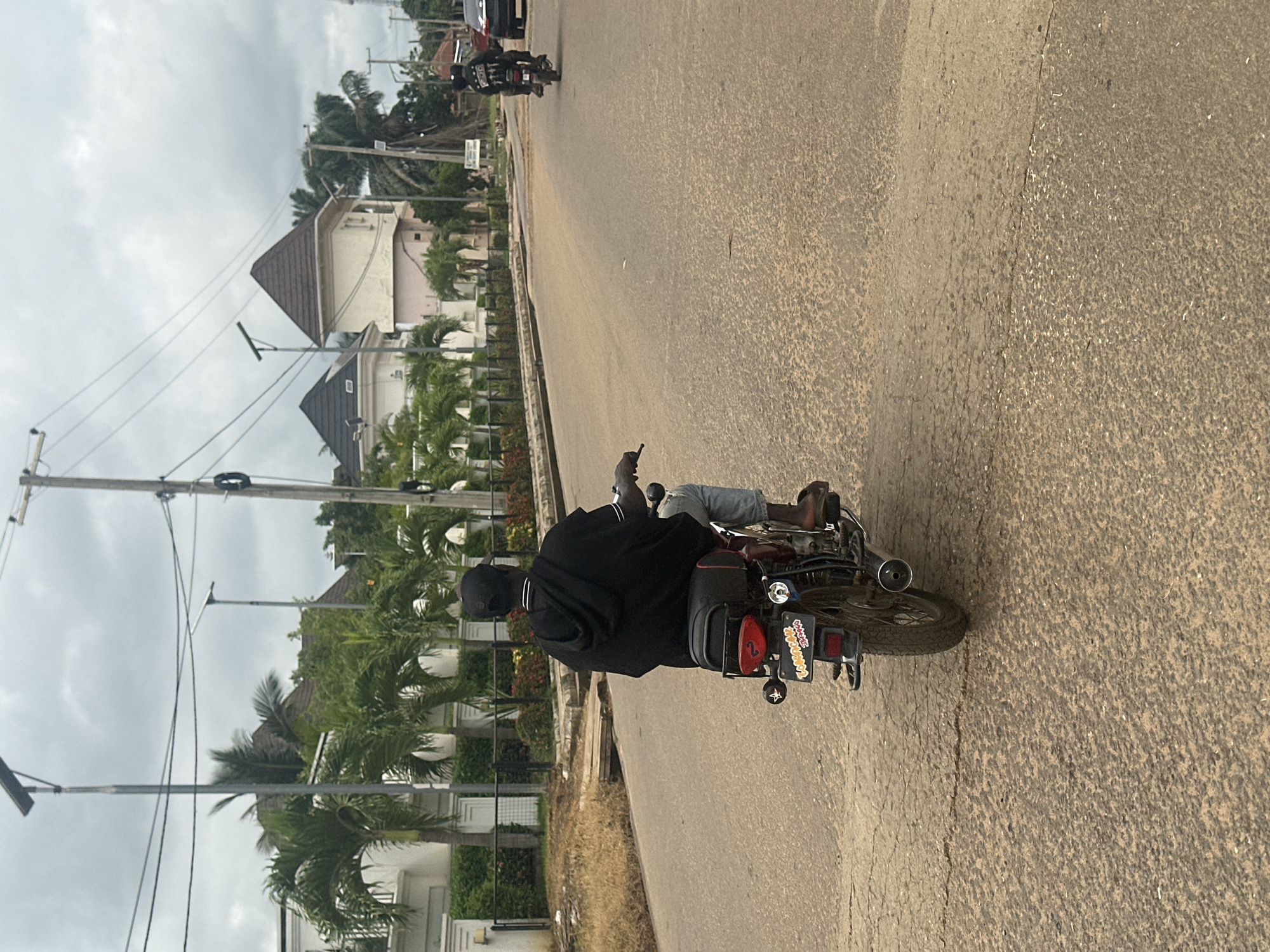 A man wearing a black hooded sweatshirt and light blue jeans is riding a motorcycle down a paved road. He has a black cap on and is facing away from the camera. The motorcycle is dark colored with a red taillight and a license plate that appears to say "TANAKAJ" in yellow letters. On the left side of the road are several large, modern houses with white walls and dark roofs, surrounded by palm trees. The sky is overcast with a few visible power lines and poles. Further down the road, another motorcycle and a dark colored car are visible. The road itself is cracked and textured. This scene is located in the city of Akwanga, Nigeria.