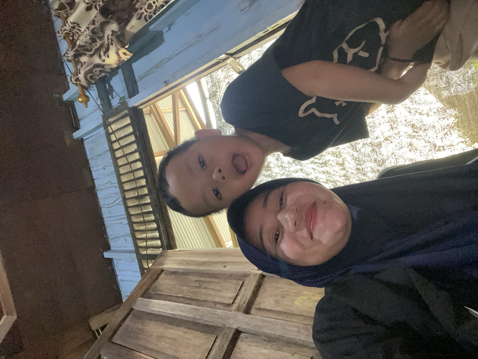 The image shows a selfie taken from a low angle, featuring a woman wearing a dark hijab and a young boy. The woman is smiling and looking at the camera, while the boy has his mouth open in an excited expression. They appear to be indoors, with wooden walls and a corrugated metal roof visible in the background. The scene suggests a casual, possibly domestic setting. The lighting is bright, indicating it might be daytime. The photo has a warm, intimate feel, capturing a moment between the two individuals.