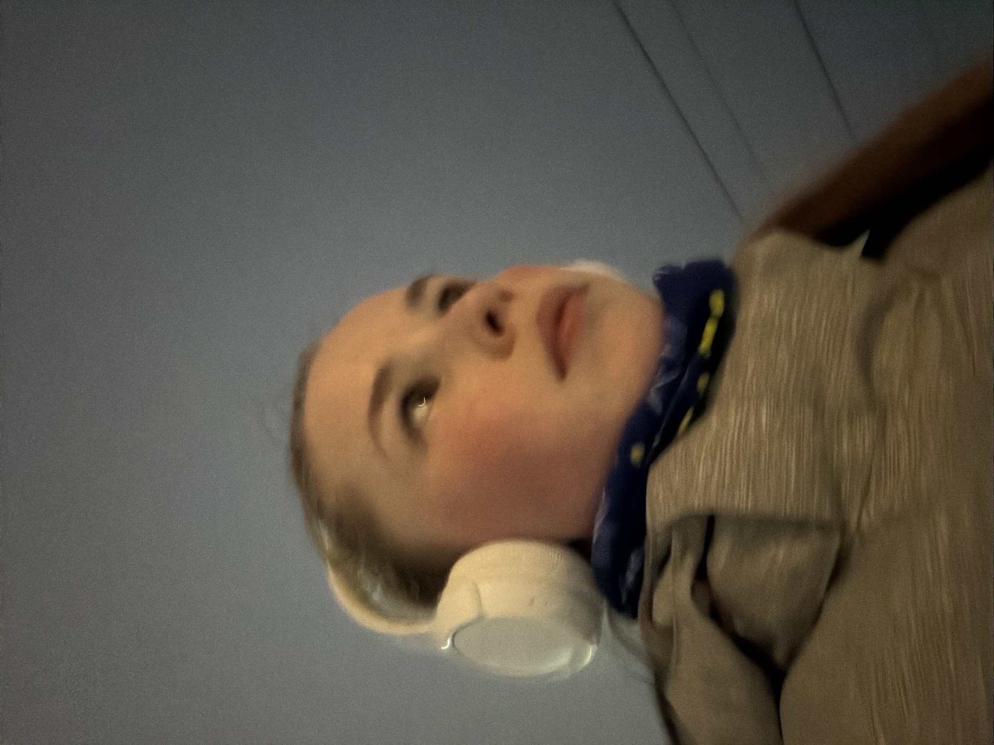 A close-up, slightly blurred, upward-angled shot depicts a person with fair skin and blondish hair wearing white headphones. The person is also wearing a blue scarf with yellow accents around their neck and a tan jacket or coat. The background is a blurred, light blue-grey, possibly indicating the sky or an indoor ceiling. The location cannot be determined definitively as Berezniki, Russia, without additional contextual information. No specific actions or interactions are occurring within the visible frame. The environment is indeterminate, suggested only by the background color.