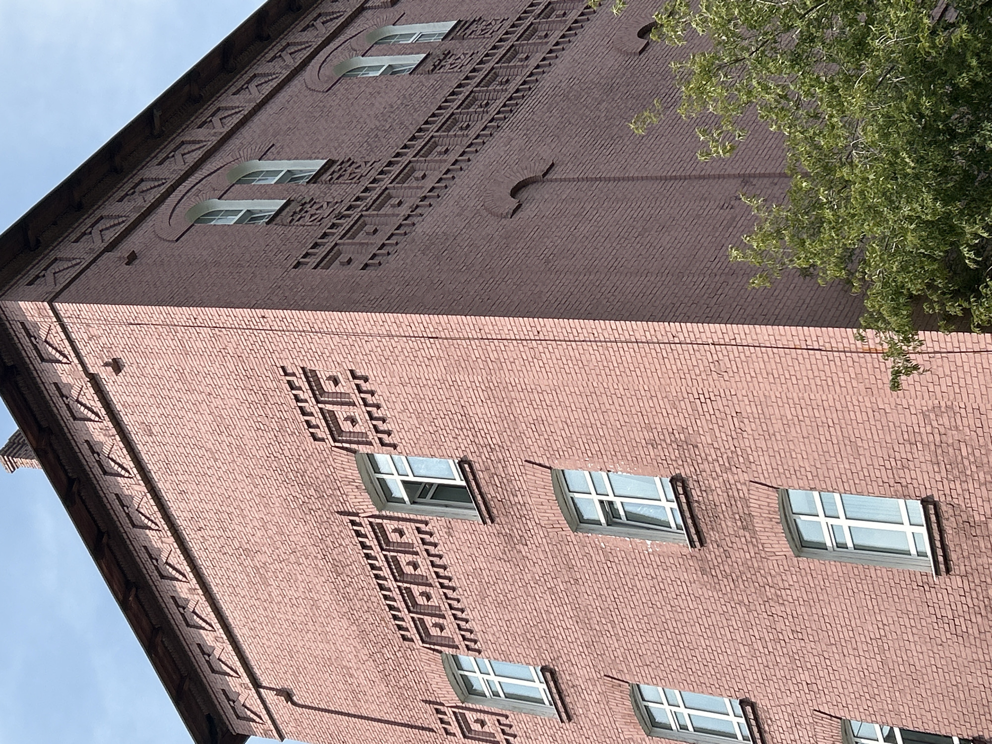 The image shows a close-up, upward view of a tall, pink brick building with a dark brown roofline. The building features multiple rows of windows with white frames, and decorative brickwork along the upper facade. The architecture suggests a historical or industrial building. A portion of a lush green tree is visible in the lower right corner, partially obscuring the building's lower levels. The sky is clear and blue. This building is the former water tower of the Tomsk railway depot, located in Tomsk, Russia.