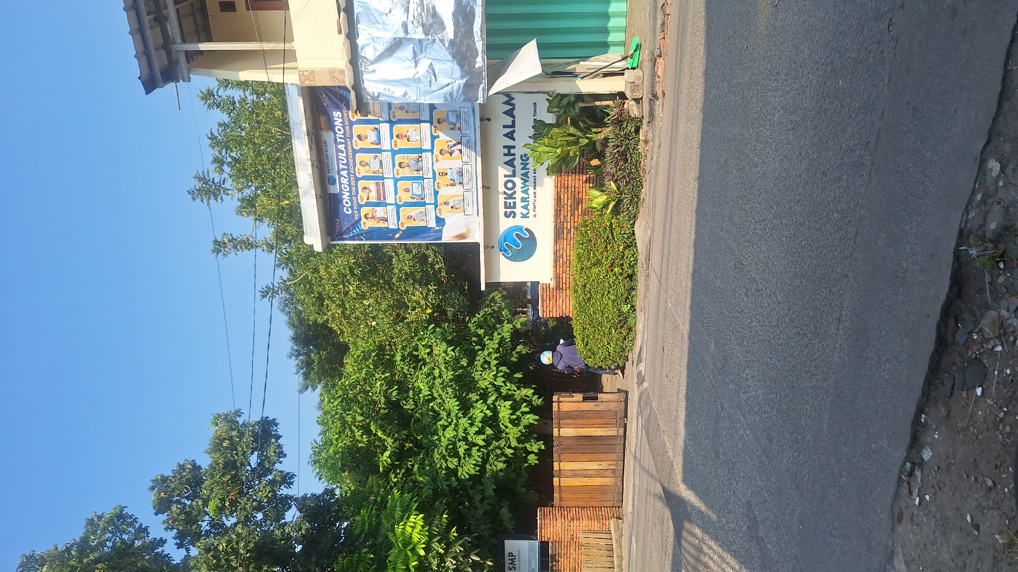The image captures a street scene in what appears to be Wadas, Indonesia. A prominent sign reads "SEKOLAH ALAM KARAWANG", accompanied by its address "JL. PINTU AIR WADAS DE". To the left of the sign, another banner displays the text "CONGRATULATIONS FOR YOUR THE BEST ACHIEVEMENT THIS MONT". A building is behind the sign, trees surround the area, and a person wearing a helmet can be seen. The street is asphalt. A sign reading "SMP" is partially visible below the wood slats.