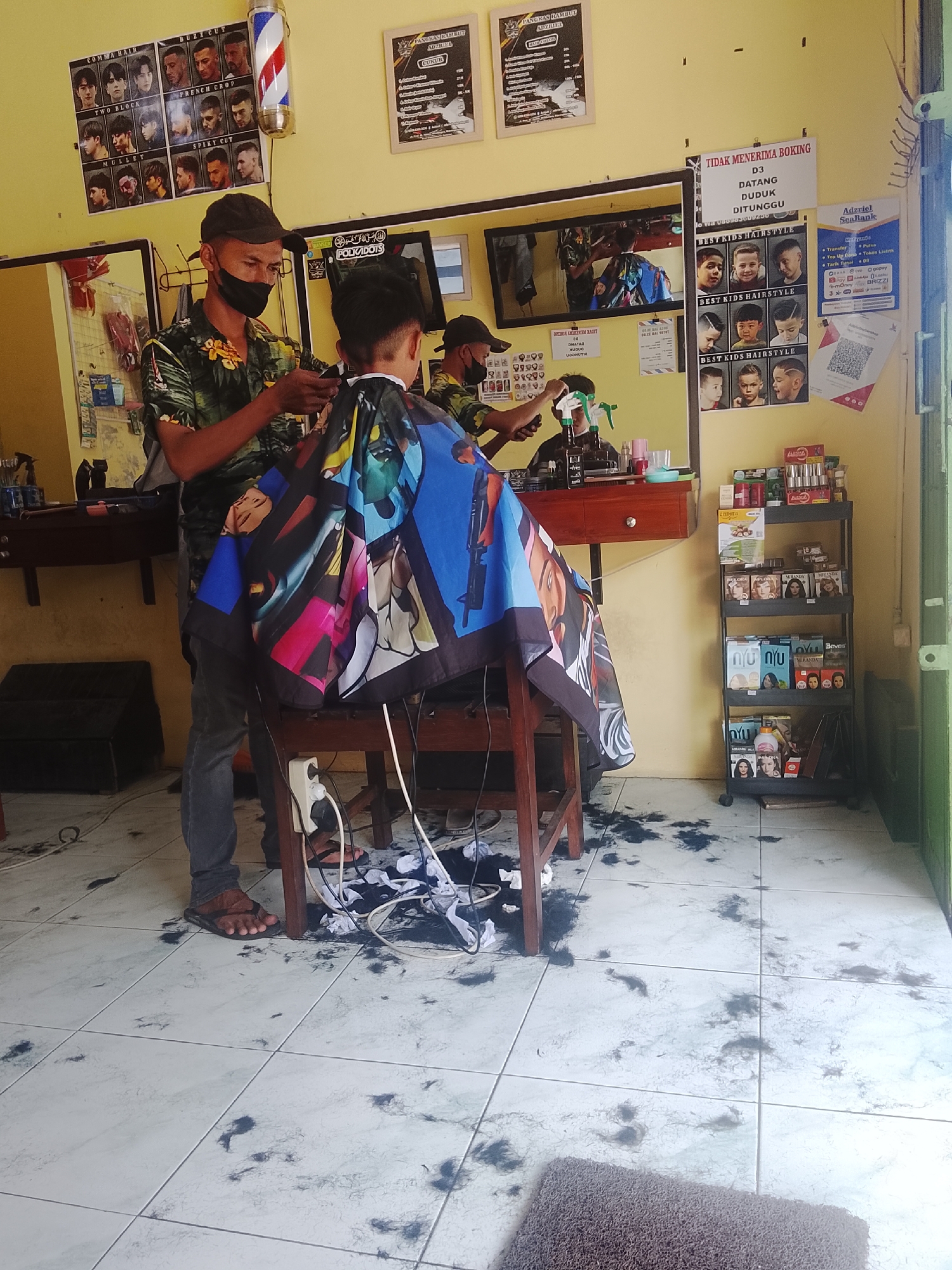 The image depicts the interior of a barbershop, likely located in Karangklesem, Indonesia. A barber, wearing a camouflage-patterned shirt and a dark cap, is in the process of cutting the hair of a young client. The barber is holding hair clippers and is focused on the client's head, who is seated and draped with a colorful cape featuring cartoonish characters. The floor of the barbershop is covered in scattered hair clippings, indicative of recent haircuts.
In the background, various elements contribute to the barbershop's atmosphere. A mirror reflects the scene, showing another barber, or possibly the same one, in action. Posters displaying different hairstyles are mounted on the walls, serving as a visual menu for customers. A barber pole, a classic symbol of barbershops, stands near the ceiling. On the right side of the image, a multi-tiered shelf unit is stocked with various hair products and personal care items. A small sign on the wall near the shelf reads "TIDAK MENERIMA BOKING," which translates to "NO BOOKING ACCEPTED." Another sign below it says "DATANG DUDUK DITUNGGU," meaning "COME, SIT, WAIT." A flyer for "Adzriel SeaBank" is also visible on the wall, suggesting a payment or financial service partnership. The overall environment appears functional and typical of a local barbershop.
