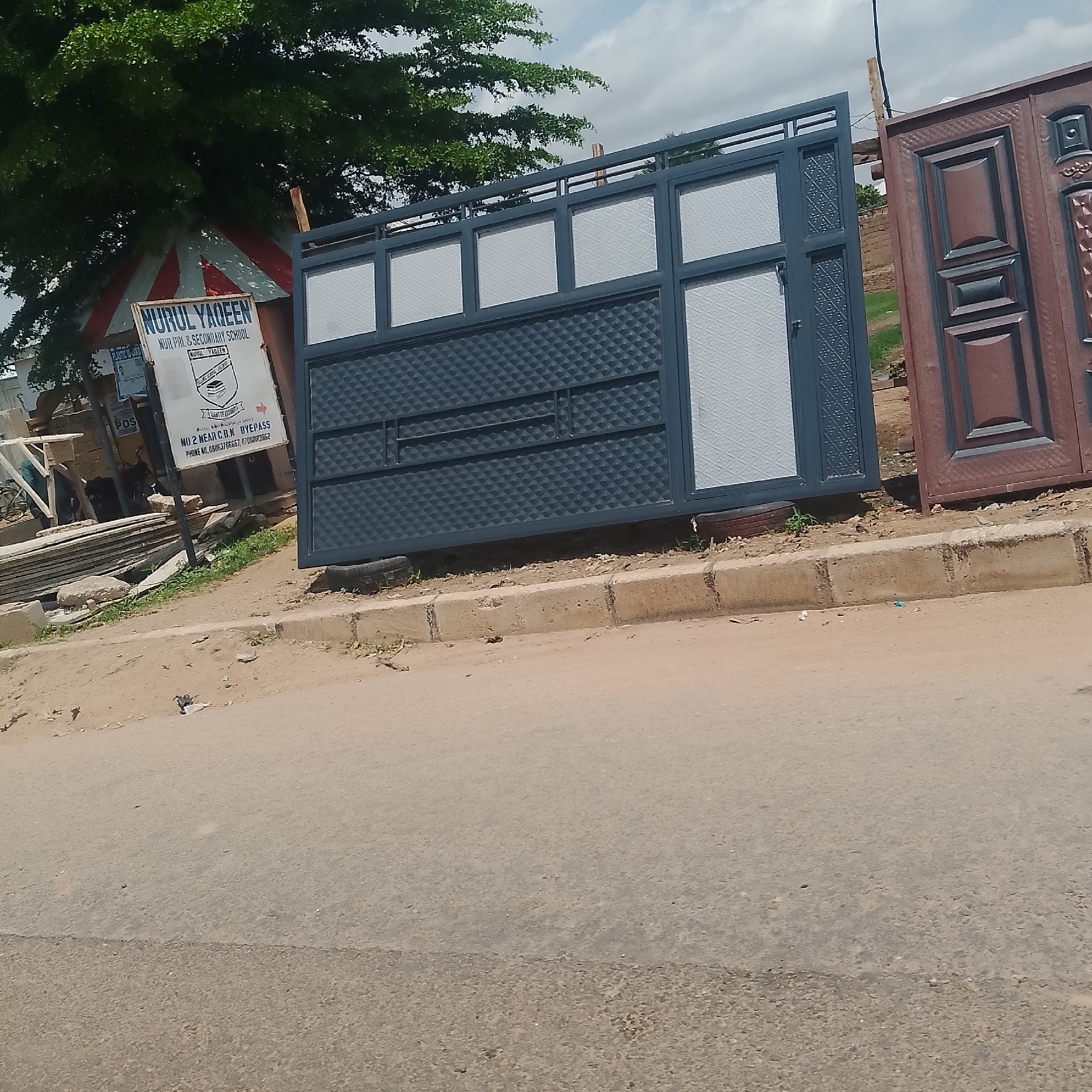 The image captures an outdoor scene in Jalingo, Nigeria, featuring a dark grey metal gate with textured diamond patterns and rectangular panels. To the right of this gate, a portion of a reddish-brown metal gate with decorative panels is visible. In the background, a sign for "NURUL YAQEEN NUR. PRY & SECONDARY SCHOOL" is displayed, along with contact information. Adjacent to the sign, a striped red and white tent is set up, and to the left, a stack of grey flat objects, possibly building materials, are piled. A tree with lush green foliage overhangs the scene from the top left. The ground is a dusty, uneven surface with scattered debris, bordered by a concrete curb. The sky above is partly cloudy.