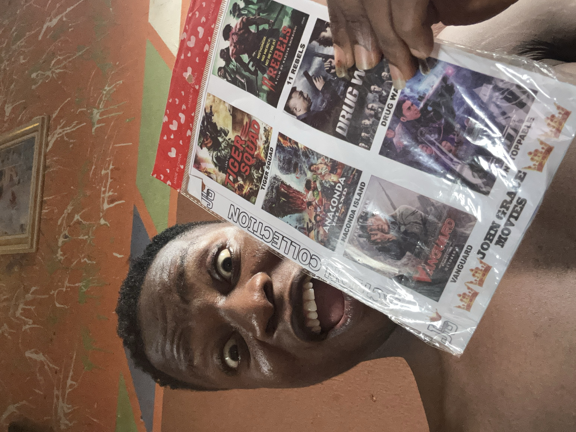 A close-up, rotated shot shows a man holding a movie collection package. The collection includes titles like "11 Rebels," "Drug War," "Tiger Squad," "Anaconda Island," and "Vanguard" featuring Jackie Chan. The collection is labeled "John Grace Movies." The package has "Made in China" printed on it and a small sticker with "JP" on the bottom corner. The man's face is partially obscured by the movie package, revealing his open mouth and teeth. The background features a wall painted with orange and green patterns and a framed picture.