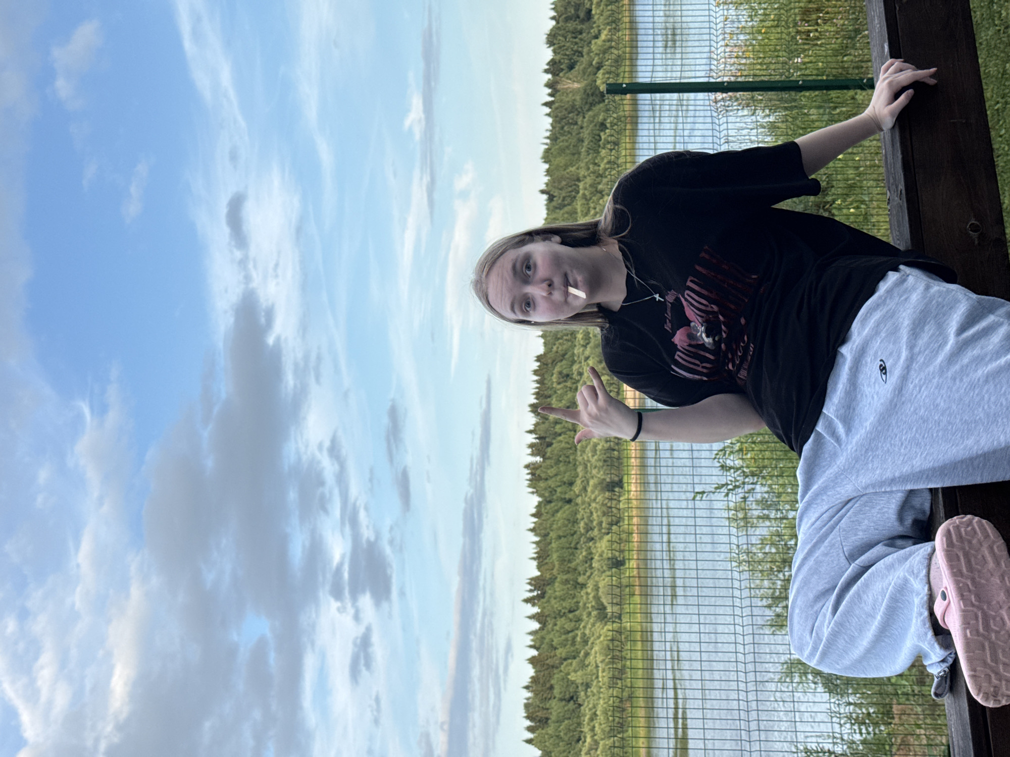 A young Caucasian female is seated on a wooden railing outdoors, with a body of water and a treeline in the background. She has blonde hair and is wearing a black t-shirt and light gray sweatpants. A white object, possibly a cigarette or a toothpick, is in her mouth. Her right hand is raised, making a hand gesture. The environment appears to be a park or recreational area, possibly near a river, with a fence separating the seating area from the water. The sky is partly cloudy. The location is identified as Ryabinino, Russia.