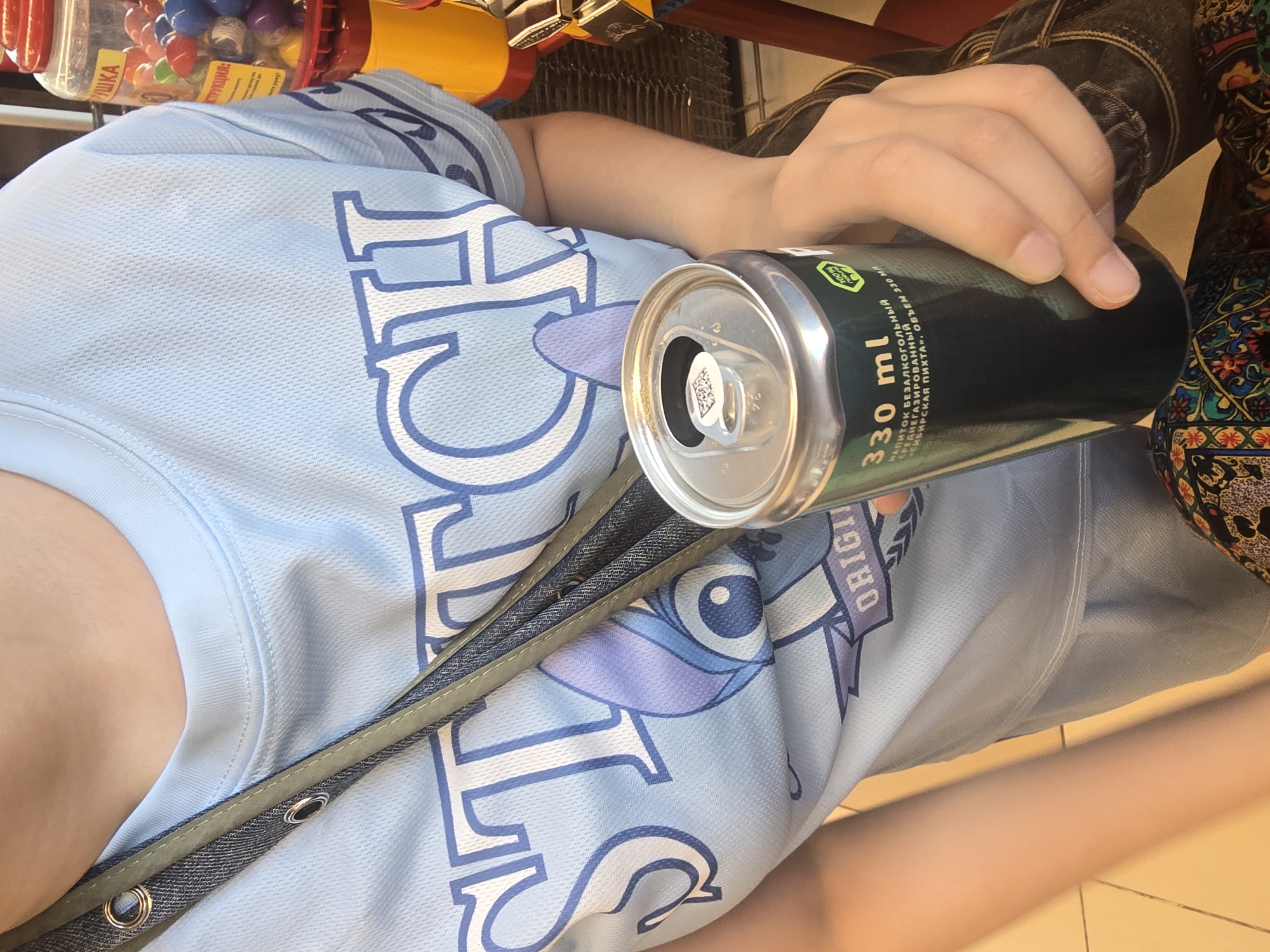 A person is holding a 330 ml can of a non-alcoholic drink, possibly in a store in Ulan-Ude, Russia. The can is dark green and has some text in Russian, including "напиток безалкогольный" (non-alcoholic drink) and "объем 330 мл" (volume 330 ml). The person is wearing a light blue t-shirt with a Stitch graphic on it, and a dark denim jacket. In the background, there is a yellow and red gumball machine filled with colorful gumballs. The person's hand is holding the can, and their arm is visible. The flooring appears to be tiled.