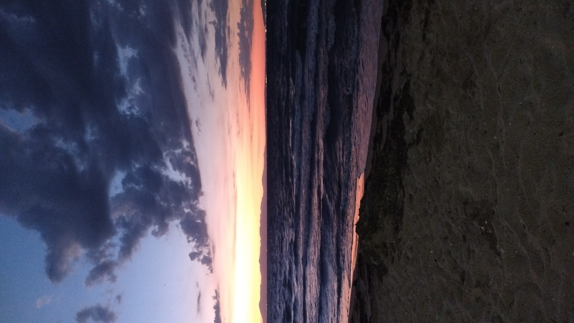 The image depicts a sunset over the Aegean Sea in Perea, Greece. The foreground is dominated by a dark, sandy beach with some scattered debris. The sea stretches out into the mid-ground, with gentle waves reflecting the colors of the sky. In the distance, a range of mountains forms the horizon, with faint lights suggesting a distant settlement. The sky transitions from a deep blue at the top to vibrant hues of orange and pink near the horizon, punctuated by dramatic, dark clouds. No people or animals are visible in the scene.