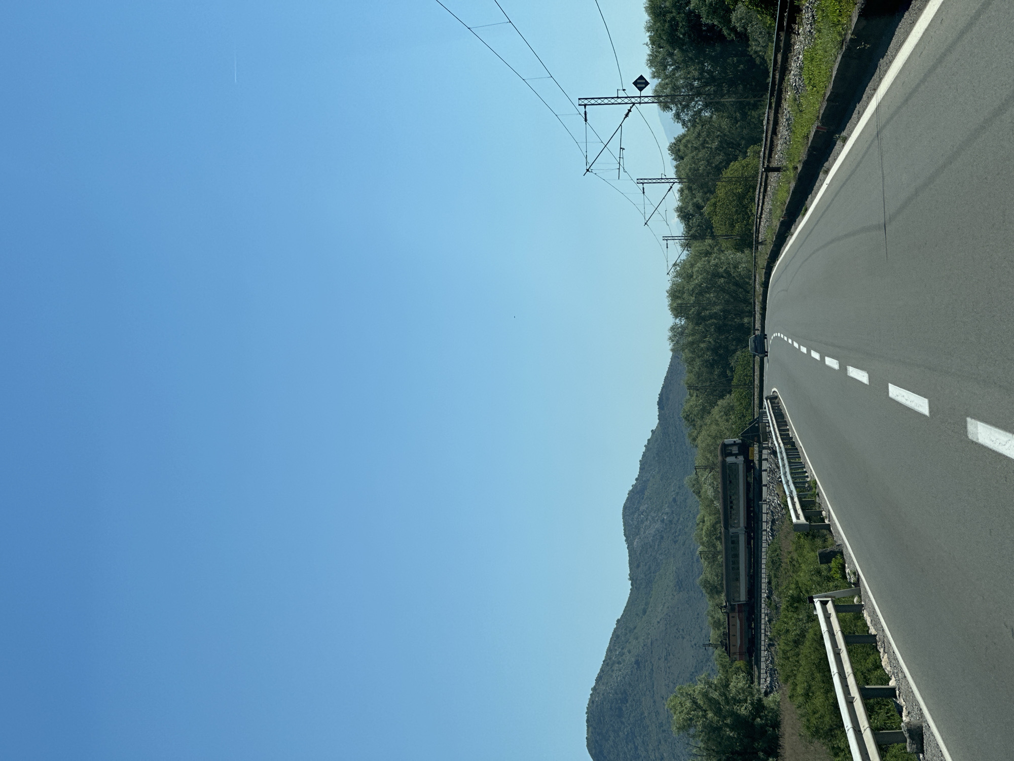 An image showing a stretch of road in Virpazar, Montenegro, next to a train track. The road has white dashed lane markings and is bordered by a guardrail. Adjacent to the road is a railway line with overhead electrical infrastructure. A train is visible on the tracks, set against a backdrop of green hills and a clear blue sky. The image is oriented in a portrait style, with the horizon aligned along the top edge of the frame.