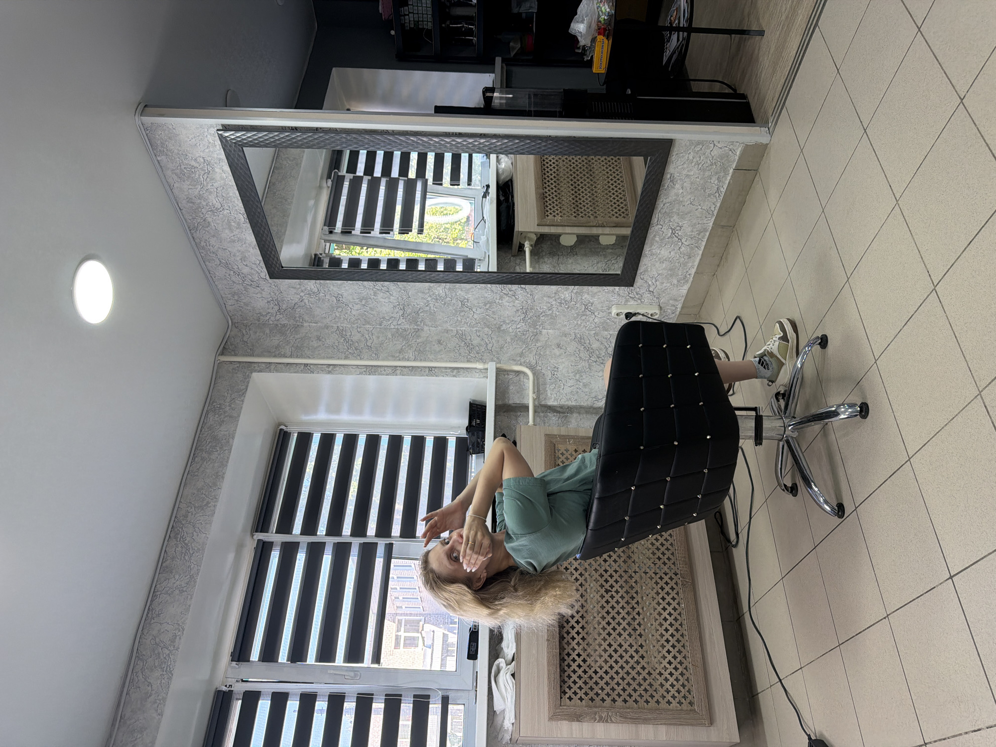 This image captures the interior of what appears to be a beauty salon or hair studio in Chaikovsky, Russia.
The scene shows a woman with blonde, wavy hair, wearing a light green t-shirt, seated in a black, quilted salon chair with a chrome base and five-spoke wheeled legs. She is turned slightly towards the viewer, with her hands raised to her face, partially covering her mouth, and her eyes wide open, suggesting a gesture of surprise or a playful expression. She is wearing white Adidas socks and multi-colored athletic shoes (beige, white, and a darker accent).
The room features light beige square ceramic floor tiles with dark grey grout. The walls are covered in a greyish wallpaper with a mottled, textured, abstract pattern. A white coving runs along the top edge of the walls, meeting a plain white ceiling from which a single circular LED light fixture emits illumination on the right side.
On the left side of the frame, a large multi-pane window is fitted with horizontal zebra blinds, alternating between opaque dark stripes and translucent lighter stripes. Through the window, glimpses of exterior buildings and some greenery are visible. Below the window is a white windowsill and a light-colored wooden or laminate radiator cover with a decorative, geometric lattice pattern. On the windowsill, a stack of folded white towels is visible to the left, a black hairdryer with its cable lies in the center, and a black basket or container is on the right. A white pipe runs horizontally along the wall below the window sill, connecting to a vertical section.
To the right, a large, rectangular mirror with a dark, textured frame hangs vertically on the wallpapered wall. The mirror reflects the window with its blinds, the exterior view, and the radiator cover. Further to the right, through an open doorway, a darker-colored wall is visible, along with a black shelving unit containing various items, a small round black table with a yellow box (possibly sweets) and other objects, and a black stand-up unit, likely additional salon equipment. A black power cable runs across the floor from under the radiator cover towards the left side of the frame. Another black cable runs from near the base of the chair towards a white electrical outlet on the wall.