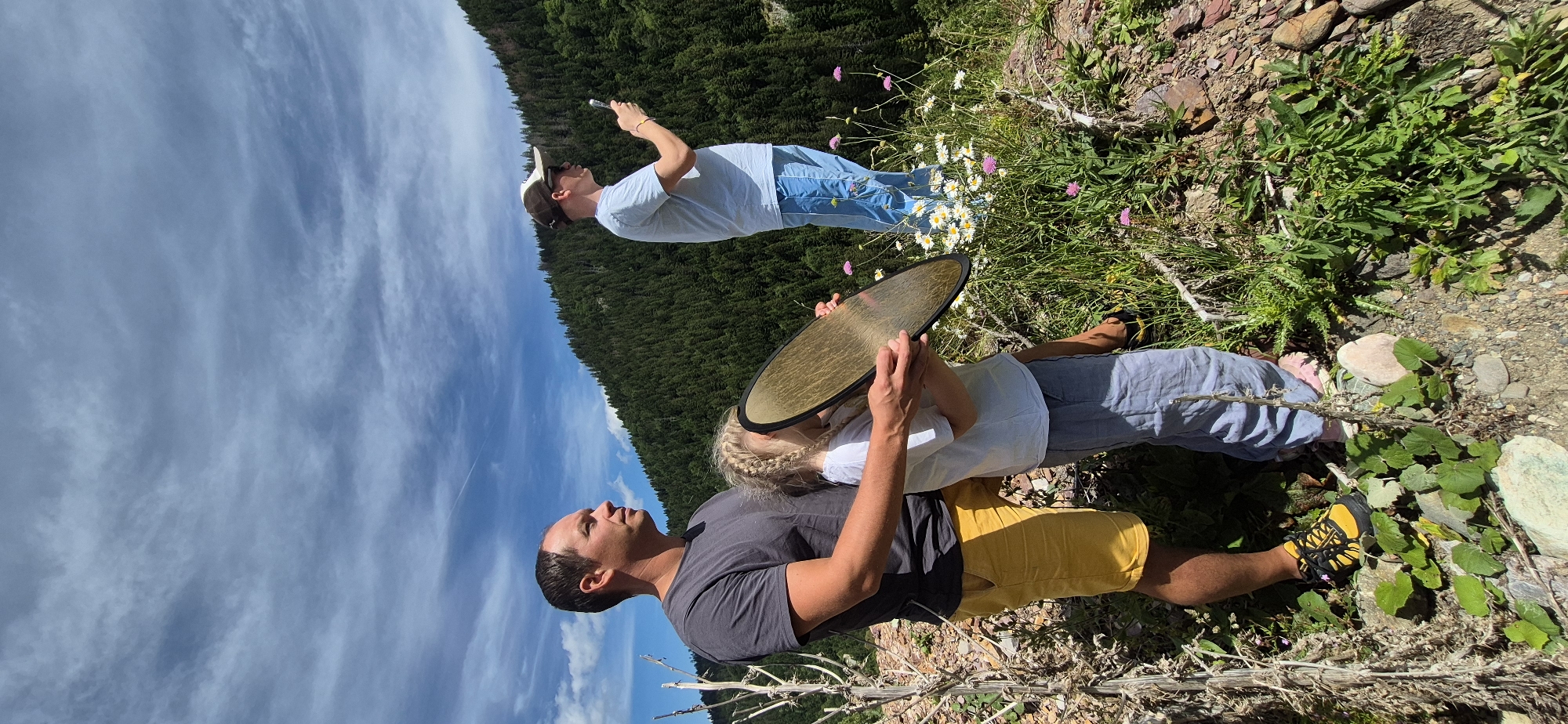 Three individuals are positioned on a mountain slope overlooking a dense forest under a partially cloudy sky. In the foreground, a man in a grey t-shirt and yellow shorts stands to the left, embracing a young child who is holding a reflective disc. The child wears a white t-shirt and grey pants. To the right and further back, a man in a light blue t-shirt, blue shorts, a baseball cap, and sunglasses holds a smartphone, appearing to take a picture. The environment consists of rocky terrain with sparse vegetation, including green plants and small white and pink wildflowers. The background is dominated by a steep mountainside covered in dark green evergreen trees, reaching towards a blue sky with scattered white clouds. There is no indication of the city of Sedlačka gora, Montenegro in the image.
