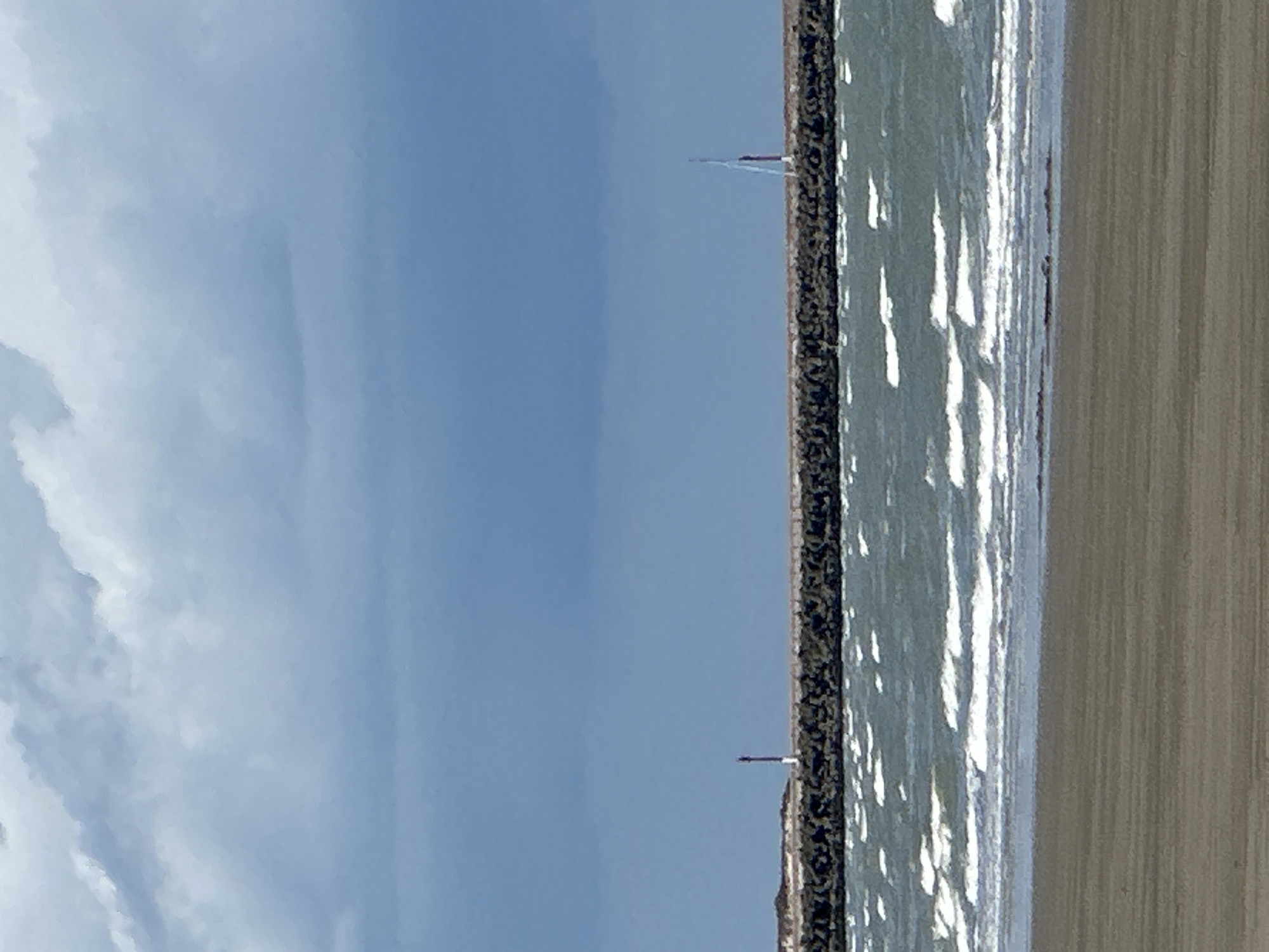 The image shows a view of the sea and a breakwater at Dunkirk, France. The sky is partly cloudy with dramatic, dark clouds in the distance and lighter clouds closer to the horizon. The sea is a greenish-blue color with white-capped waves. A sandy beach is visible in the foreground. The breakwater is made of large, dark rocks and stretches across the middle ground of the image. On the breakwater, to the left, is a small lighthouse with a red top and white pole. Further to the right on the breakwater, a white sailboat with a red stripe on its hull is visible. The overall scene suggests a somewhat moody or unsettled weather day at the coast.