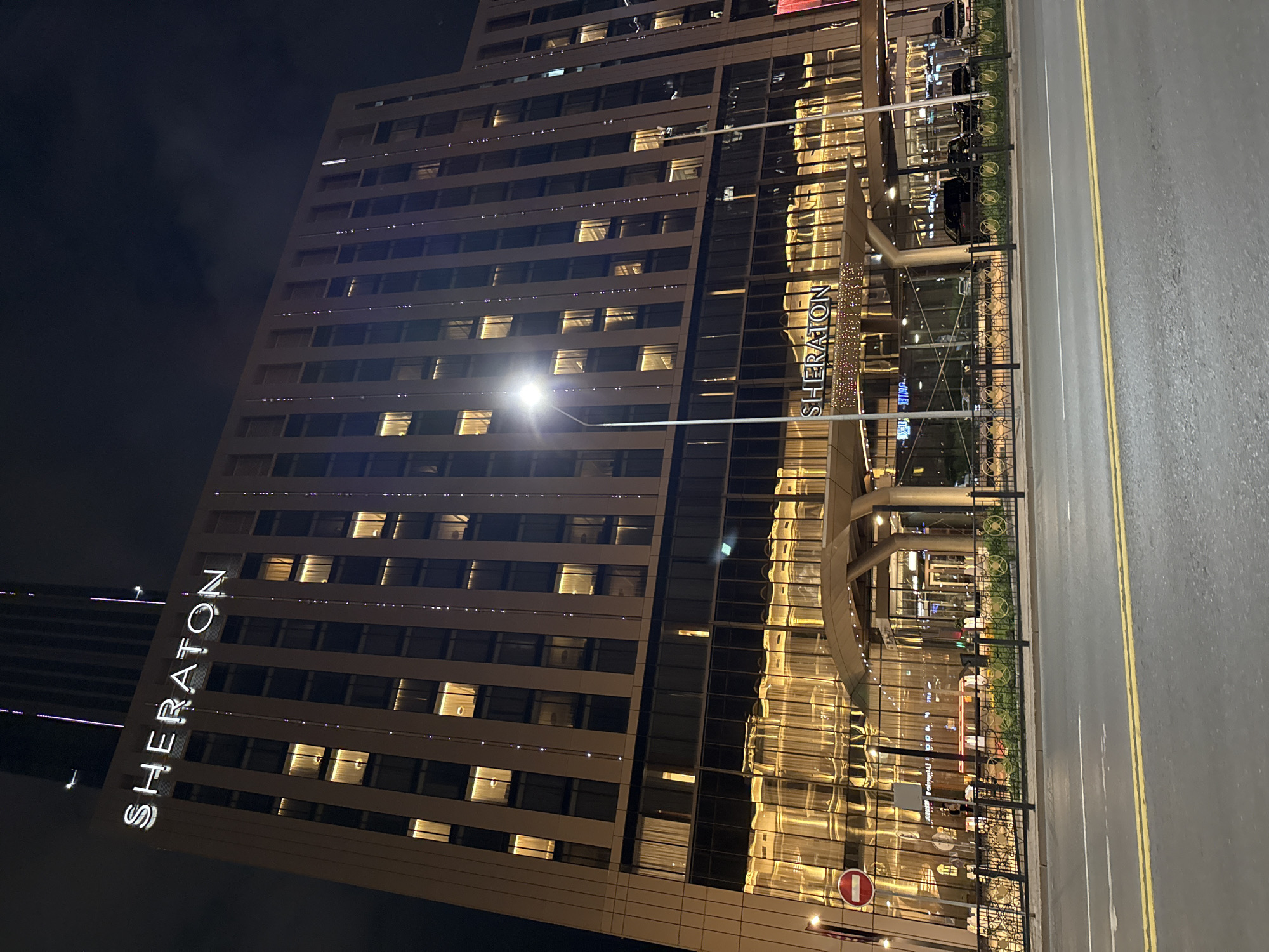 This media file captures a nighttime exterior view of the Sheraton Hotel in Astana, Kazakhstan. The imposing, modern hotel building dominates the frame, with its facade featuring numerous windows reflecting the warm, golden interior lights. The building is tall and appears to be constructed with glass and light-colored materials.
Prominently displayed on the side of the hotel is the "SHERATON" logo in large, illuminated white letters. Adjacent to the hotel, a street or roadway is visible, marked with yellow lines. A black metal fence separates the hotel entrance area from the street, with a grassy strip and some decorative elements behind the fence.
The scene is illuminated by the hotel's internal lighting and streetlights, creating a well-lit but dark nighttime atmosphere. The sky is dark, suggesting it is late at night or early morning. There are no visible people clearly identifiable, but reflections in the windows hint at some activity inside. A red and white circular sign, likely a traffic or informational sign, is visible near the entrance. The overall impression is one of a well-lit, upscale urban hotel at night.