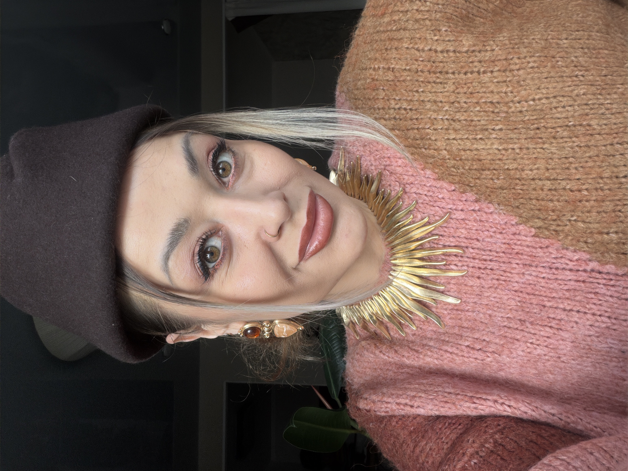 A woman is depicted from the chest up, wearing a brown beret, gold-toned necklace resembling a sunburst, and a multi-toned sweater with pink and brown hues. She is wearing earrings with two amber-like jewels and has light blonde hair with darker roots. Her makeup includes lipstick and eye shadow with a shimmer finish. A dark-colored wall and a leafy green plant are slightly visible in the background. There is no information in the image to confirm the location.