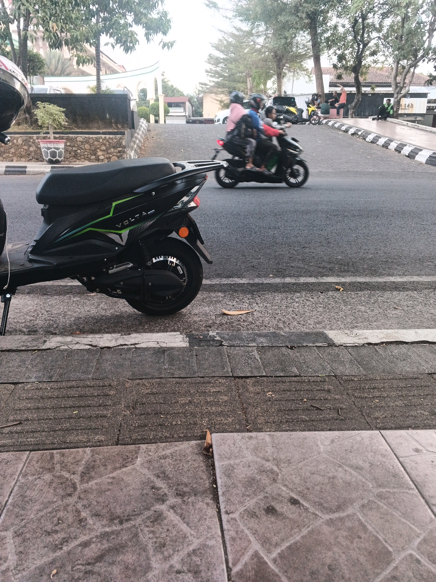 In Sidomulyo, Indonesia, a street scene unfolds with a focus on a black electric scooter in the foreground, parked on a textured gray and brown sidewalk. The scooter, branded with "VOLTA" in green and white, is angled towards the right of the frame, revealing its rear wheel and electric motor. Behind the scooter, the asphalt road features a black and white striped curb. Several motorcycles and people are visible on the road in the middle ground. One motorcycle, carrying three individuals, is in motion, creating a slight blur. Another motorcycle is parked. People are standing and sitting along the side of the road, with one person in a green shirt seated and looking towards the camera. In the background, trees and buildings with white architectural elements are present, suggesting a mixed urban and residential environment. The sky is overcast, contributing to the soft lighting of the scene.