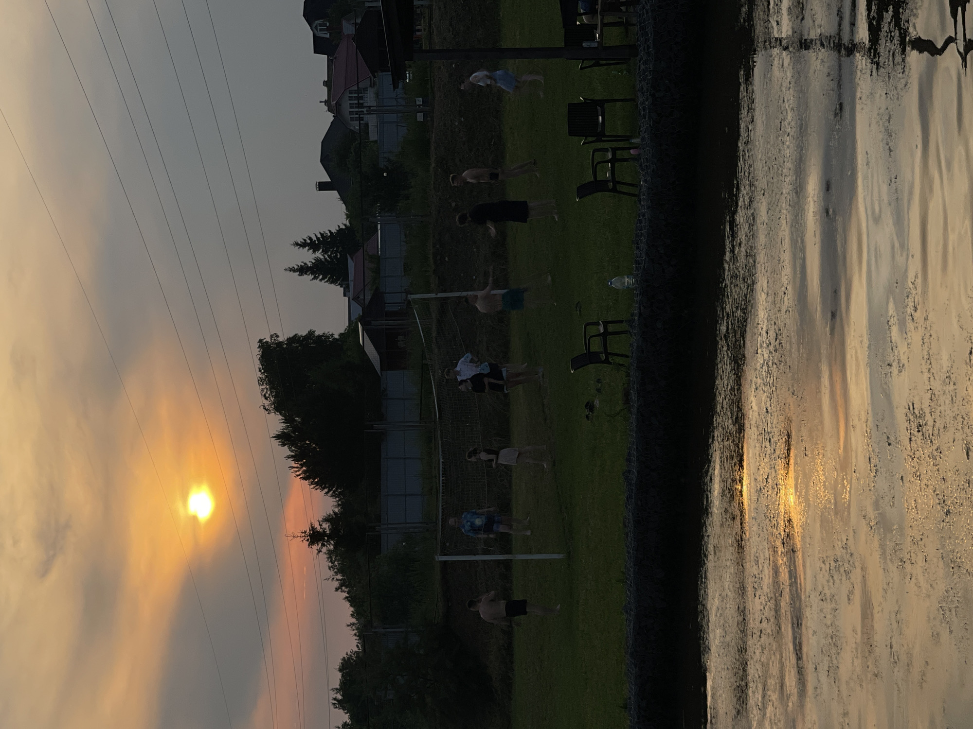 The image depicts an outdoor scene at dusk, likely in Novoseltsevo, Russia, featuring a group of people playing volleyball on a grassy field. The sun is low on the horizon, casting a warm, orange glow through hazy clouds, and its reflection shimmers on the surface of a body of water in the foreground. A volleyball net is set up on the field, and several individuals, appearing to be young adults, are scattered around it, engaged in or observing the game. Some are in athletic stances, suggesting active participation. In the background, trees and residential buildings are visible, silhouetted against the fading light. Power lines traverse the sky above. The overall atmosphere is relaxed and recreational.