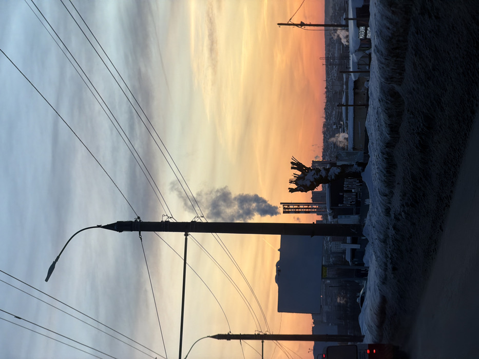An industrial cityscape under a sunset sky. Silhouetted power lines and streetlights run across the scene. In the middle ground, plumes of smoke rise from industrial chimneys. Buildings are lined up against a sky gradient of blue, yellow, and orange. Snow-covered foliage and a sign stand on the right edge, with part of a vehicle visible at the bottom.