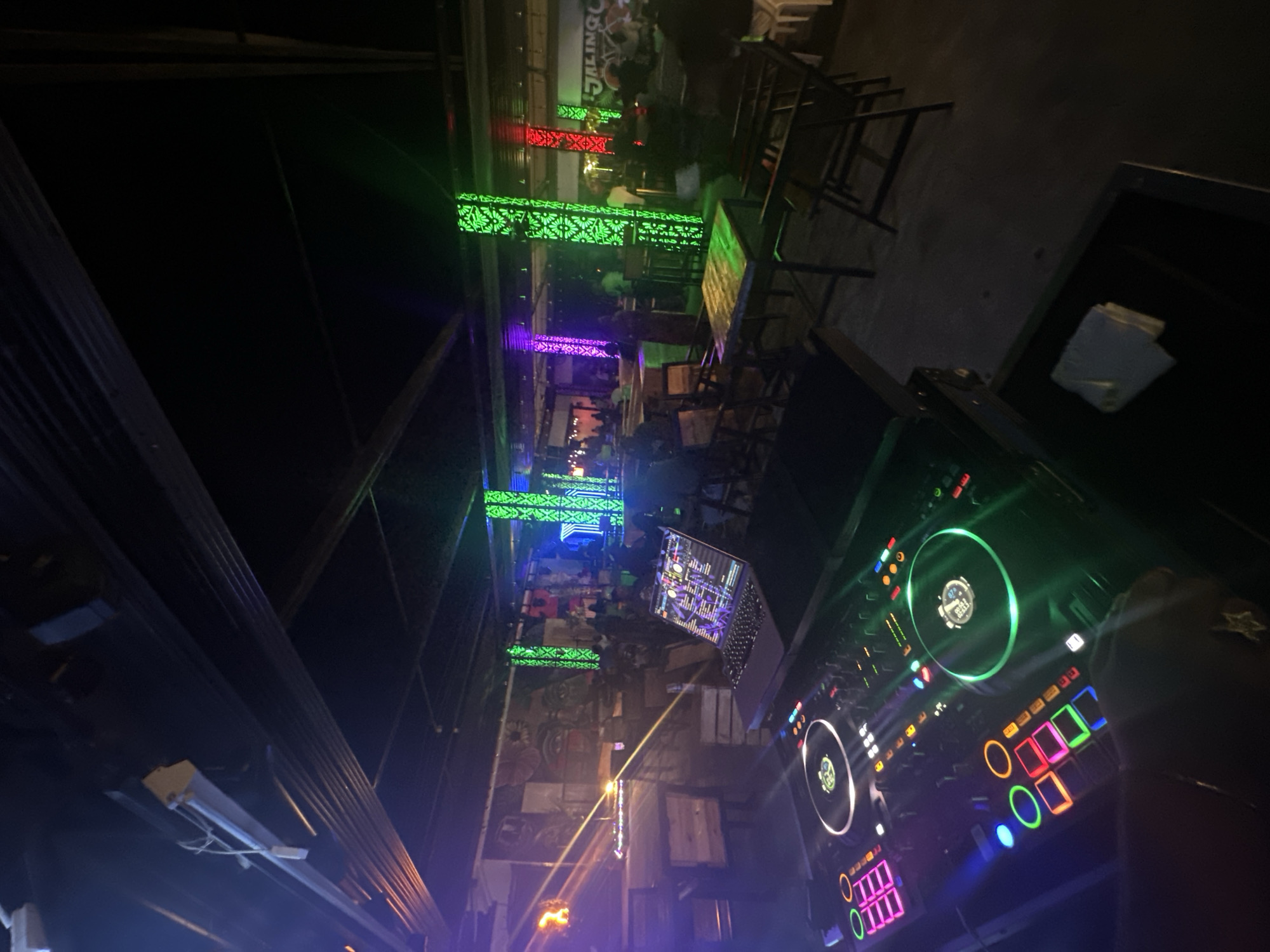 The image depicts the interior of a dimly lit establishment, likely a club or bar, in Howayi, Nigeria. In the foreground, a DJ setup is visible, featuring a laptop with a colorful display, a DJ mixer with illuminated buttons and turntables, and what appears to be a hand with a ring interacting with the equipment. The background reveals a seating area with tables and chairs, and patrons are dimly discernible. The environment is decorated with vibrant, colorful LED lighting, including green, purple, and red accents on pillars and along the ceiling. Murals or artwork adorn the walls. The overall atmosphere suggests a nightlife setting with entertainment being provided.