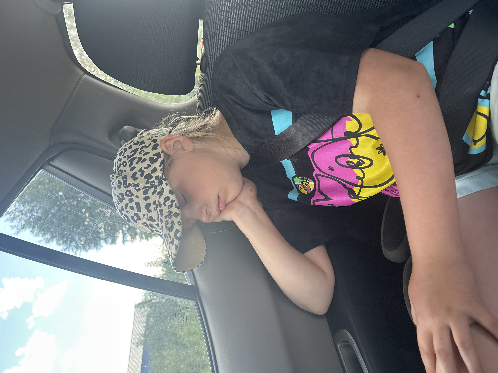 The image shows a young person sleeping in the passenger seat of a car. The person is wearing a black t-shirt with a colorful graphic design and a leopard-print baseball cap. They are leaning their head on their hand, with their eyes closed. A car seatbelt is visible, securing them in place. The background shows the interior of the car, including the window with trees and sky visible outside. The lighting suggests it is daytime. The person appears peaceful and relaxed as they sleep. The location is identified as Фоки, Russia.