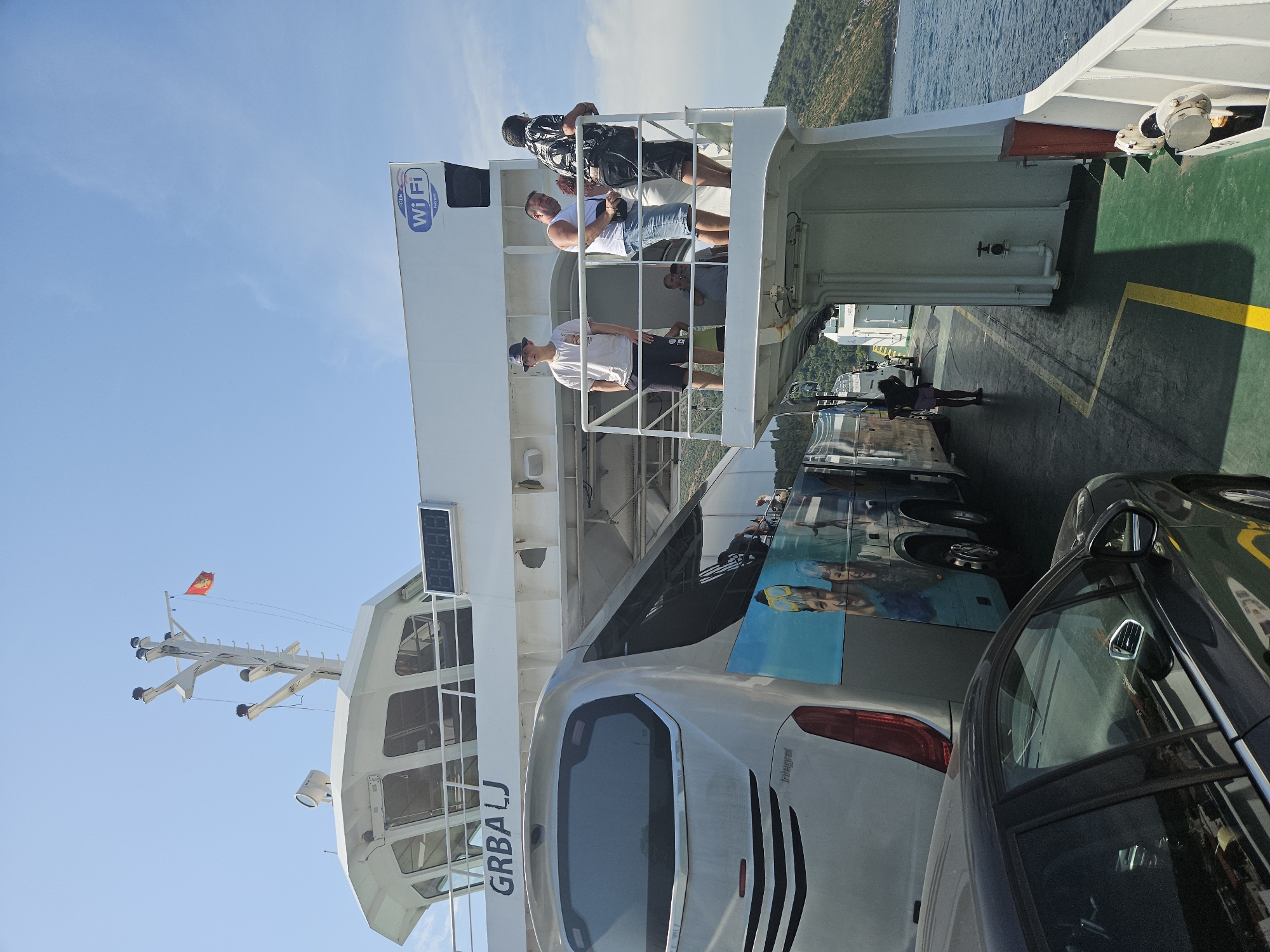 In Lepetane, Montenegro, a ferry with the name "GRBALJ" is seen docked or in the process of boarding. The ferry's deck is filled with vehicles, including a silver bus adorned with an image of people wearing scuba gear and a dark-colored car in the foreground. Several people are visible on an upper deck, including a young person wearing a hat and white t-shirt, and three men standing and looking outwards. One man wears a white tank top and denim shorts, another a black and white patterned shirt, and a third is partially visible. A "FREE Wi-Fi" sign is mounted on the side of the ferry. The sky is clear and blue, and the water is calm, with green, tree-covered hills visible in the background. The scene depicts a typical ferry crossing operation in a coastal town.