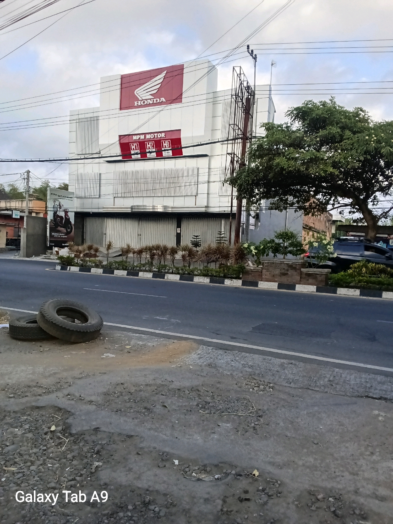 The image shows a Honda motorcycle dealership in Trenggalek, Indonesia. The building is modern with a white facade and large windows. A large red and white Honda logo is visible on the upper part of the building. Below this, a sign reads "MPM MOTOR" with smaller Honda logos. The dealership appears to be closed, with metal shutters covering the entrance.
In front of the dealership, there is a road with a sidewalk. Two used tires are lying on the side of the road in the foreground. A line of decorative plants and shrubs runs along the edge of the sidewalk. A dark-colored SUV is parked on the right side of the road, partially obscured by a tree. The sky is overcast with clouds.
The image also includes the text "Galaxy Tab A9" in the lower left corner, indicating the device used to capture the photo.