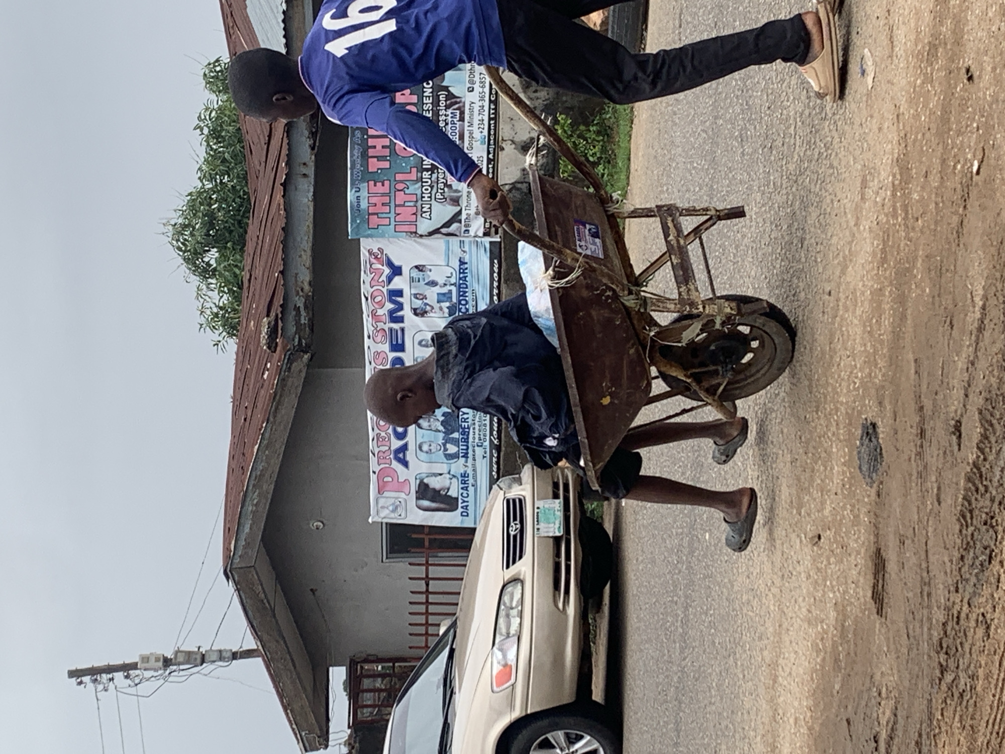 Two people move a rusty, loaded wheelbarrow on a paved road. The person closer to the wheelbarrow leans into it, while the other pushes from behind. A gold-colored sedan partially blocks the wheelbarrow's path. Advertisements for a church ministry and a school are visible on a building in the background. Electrical lines run from a pole to the side of the building. The location is inferred to be Jos, Nigeria, based on details in the advertisement.