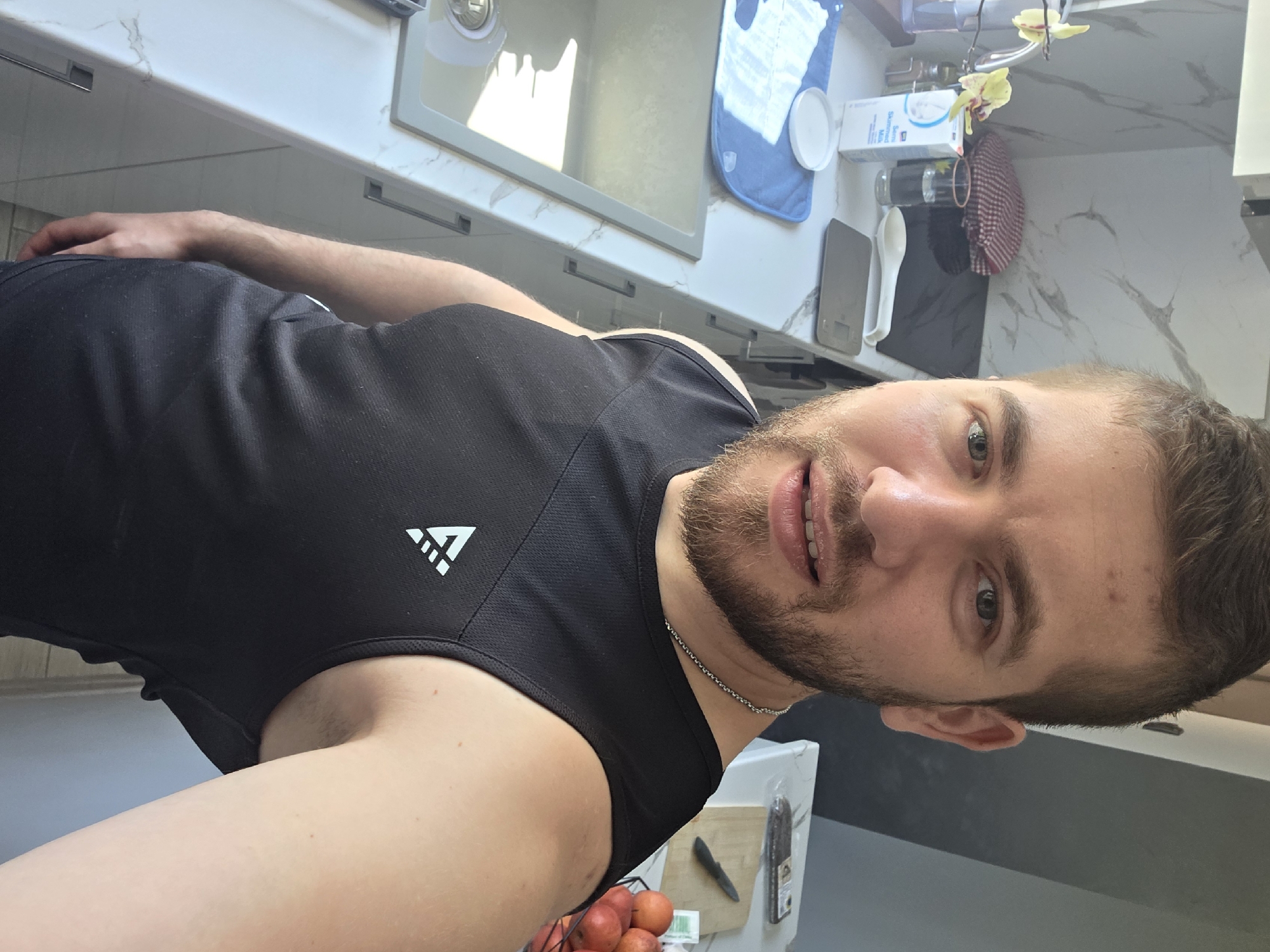 A selfie of a Caucasian male with brown hair, stubble beard, and light skin. He is wearing a black sleeveless athletic shirt with a white logo on the upper left side. He appears to be standing in a kitchen with a marble countertop, sink, kitchenware, and appliances visible in the background. A bowl of red fruits is also visible at the bottom of the frame. There is no information to connect the location to Yagodovo, Bulgaria.