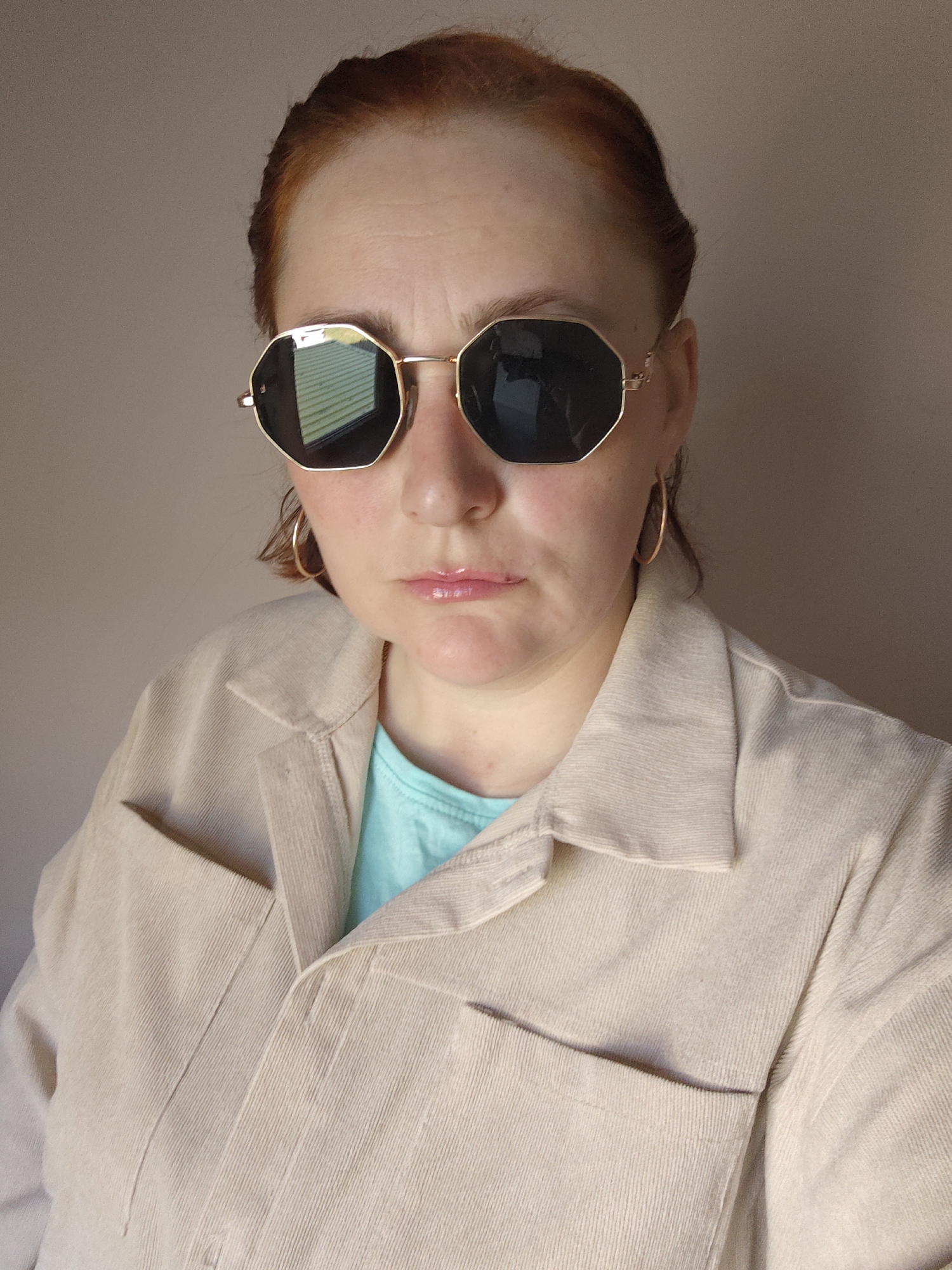 A woman with red hair is wearing octagonal sunglasses with a gold frame. She has on gold hoop earrings and a light beige corduroy jacket over a light blue shirt. The background is a solid, light beige color.