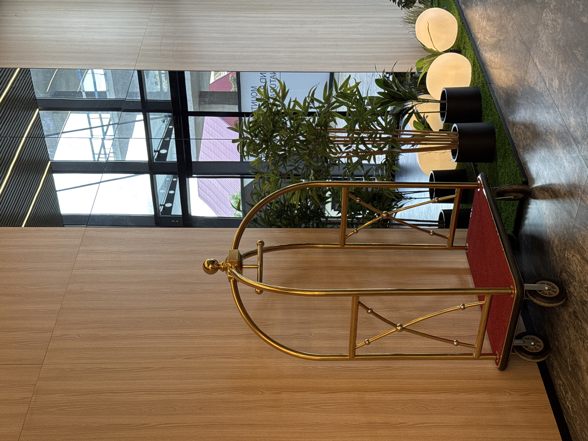 A golden luggage cart with a red carpeted base and wheels is positioned against a light wood-paneled wall in a modern indoor space. Behind the cart, a large window reveals an exterior view of buildings with purple roofs and a multi-story structure with balconies. Several potted plants, including a tall bamboo-like plant and smaller leafy plants, are arranged on a section of artificial grass. Three large, round, illuminated orbs are placed on the artificial grass next to the plants. The floor is tiled with a gray, marbled pattern. The scene appears to be in a hotel lobby or a similar public building in Batumi, Georgia.