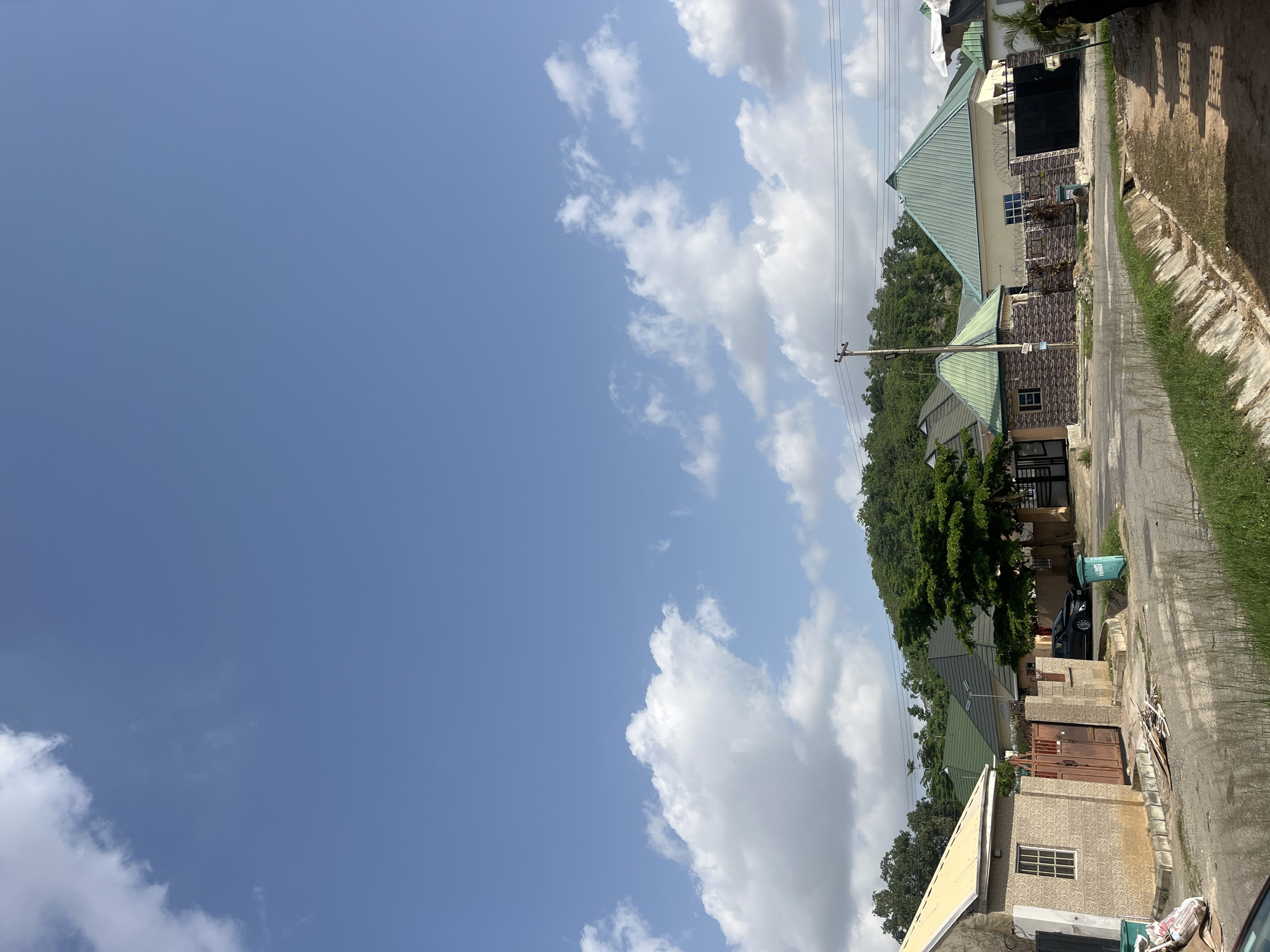 The image depicts a residential street in Abuja, Nigeria. The sky is predominantly blue with scattered white clouds. Several single-story houses with green roofs line the right side of the street, separated from the road by low walls and foliage. The houses exhibit varying architectural styles and colors, including light beige and brown brick. A paved road runs along the right side, with a concrete drainage ditch separating the road from the houses. Green grass grows alongside the ditch. Power lines are visible against the sky. A black vehicle is parked in front of one of the houses. Trash bins are visible near some of the houses. The scene appears to be a relatively quiet residential area on a sunny day.