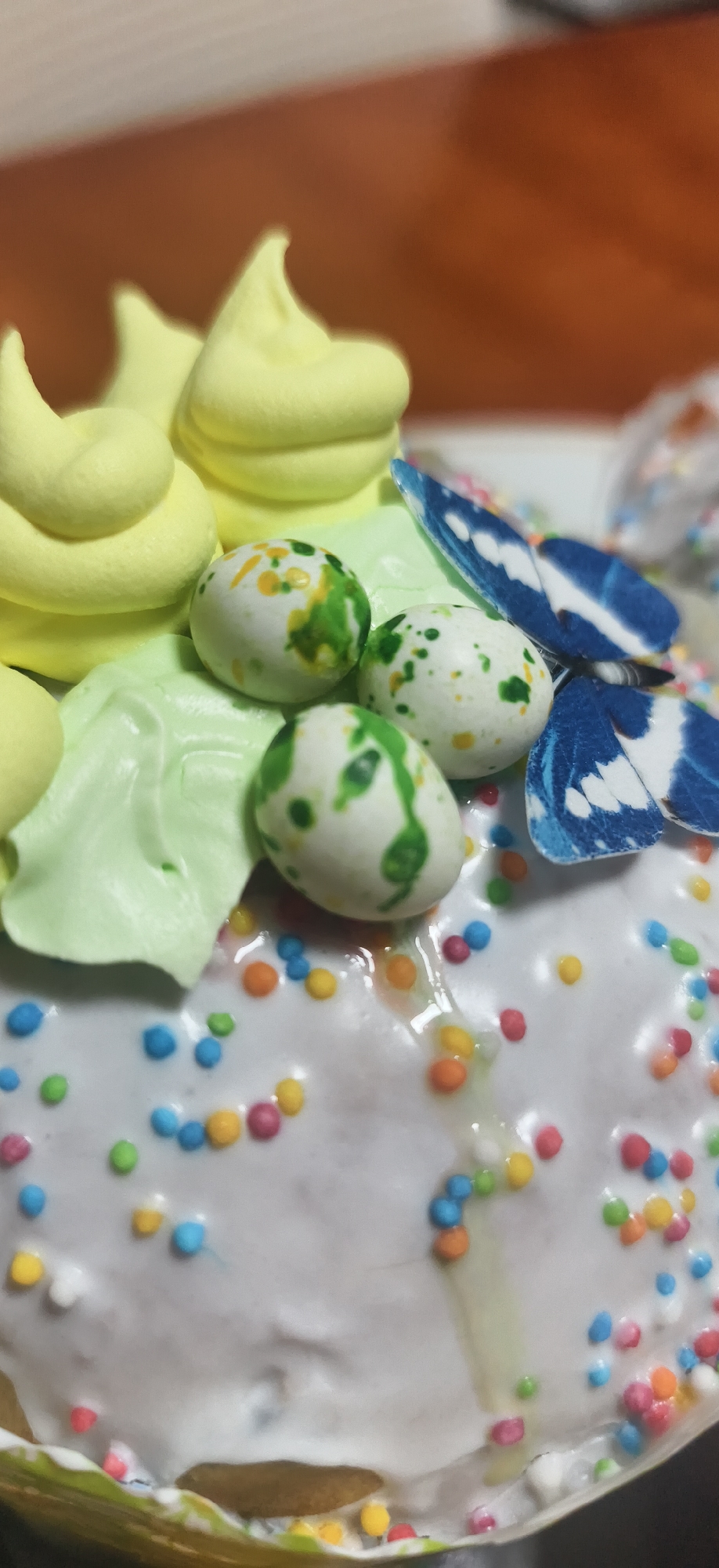 The image shows a close-up of a decorated Easter cake. The cake is covered in a white glaze and multicolored sprinkles. It is adorned with light yellow frosting swirls, light green frosting leaves, three white speckled candy eggs, and a blue butterfly decoration. The cake is likely located in Russia, given the Russian name "Фоки."