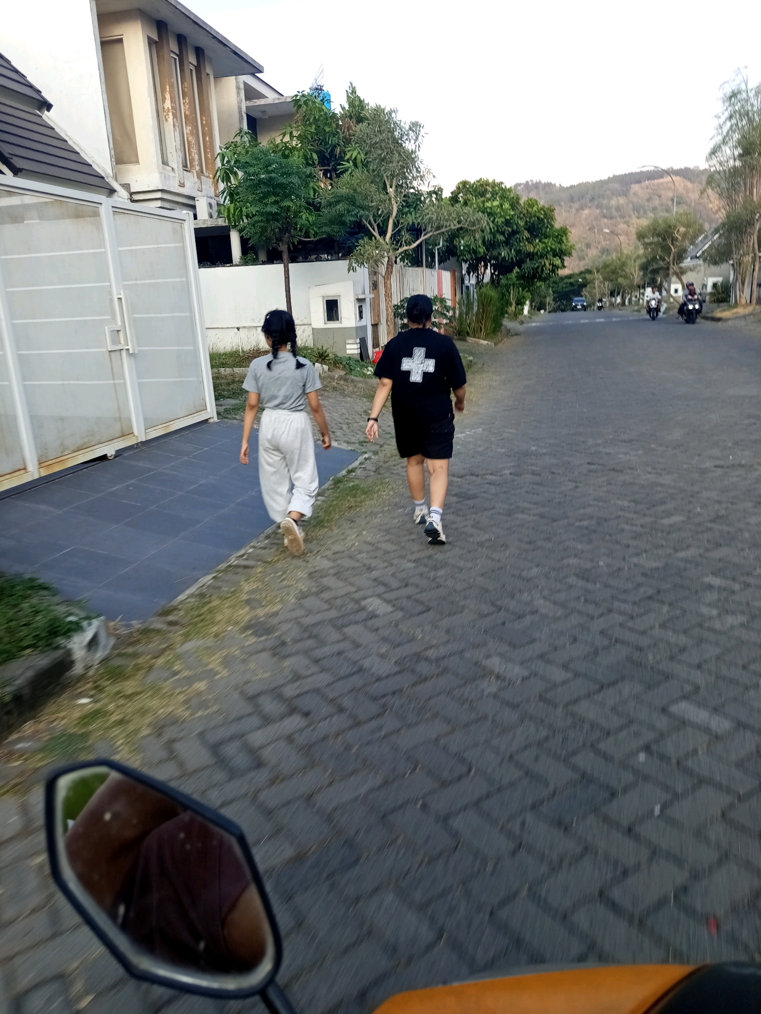 The image depicts a street scene in Sumberngepoh, Indonesia, with two individuals walking away from the camera. To the left, a person wearing a grey t-shirt and light-colored pants walks on a tiled walkway. To the right, a person in a black t-shirt with a white cross emblem and black shorts walks on a paved road. The road has a herringbone pattern of grey interlocking bricks. In the background, there are houses, trees, and a hill. Several motorcycles are also visible on the road further ahead. The photo appears to have been taken from a moving vehicle, possibly a motorcycle, as indicated by the presence of a mirror in the foreground reflecting a person's shoulder and arm. The lighting suggests it is late afternoon or early evening.