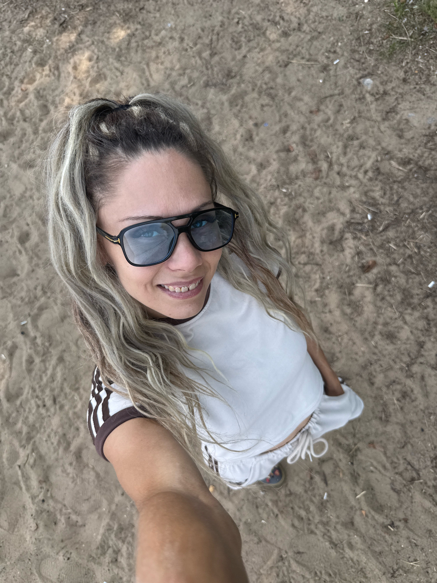A woman is standing outdoors on sandy ground. She is wearing a white crop top, white shorts, and sunglasses. Her hair is long and blonde, pulled back into a ponytail. The background is out of focus and appears to be a natural environment with some debris on the ground. The image is taken from a high angle, looking down at the woman. The location is Serenovo, Russia.