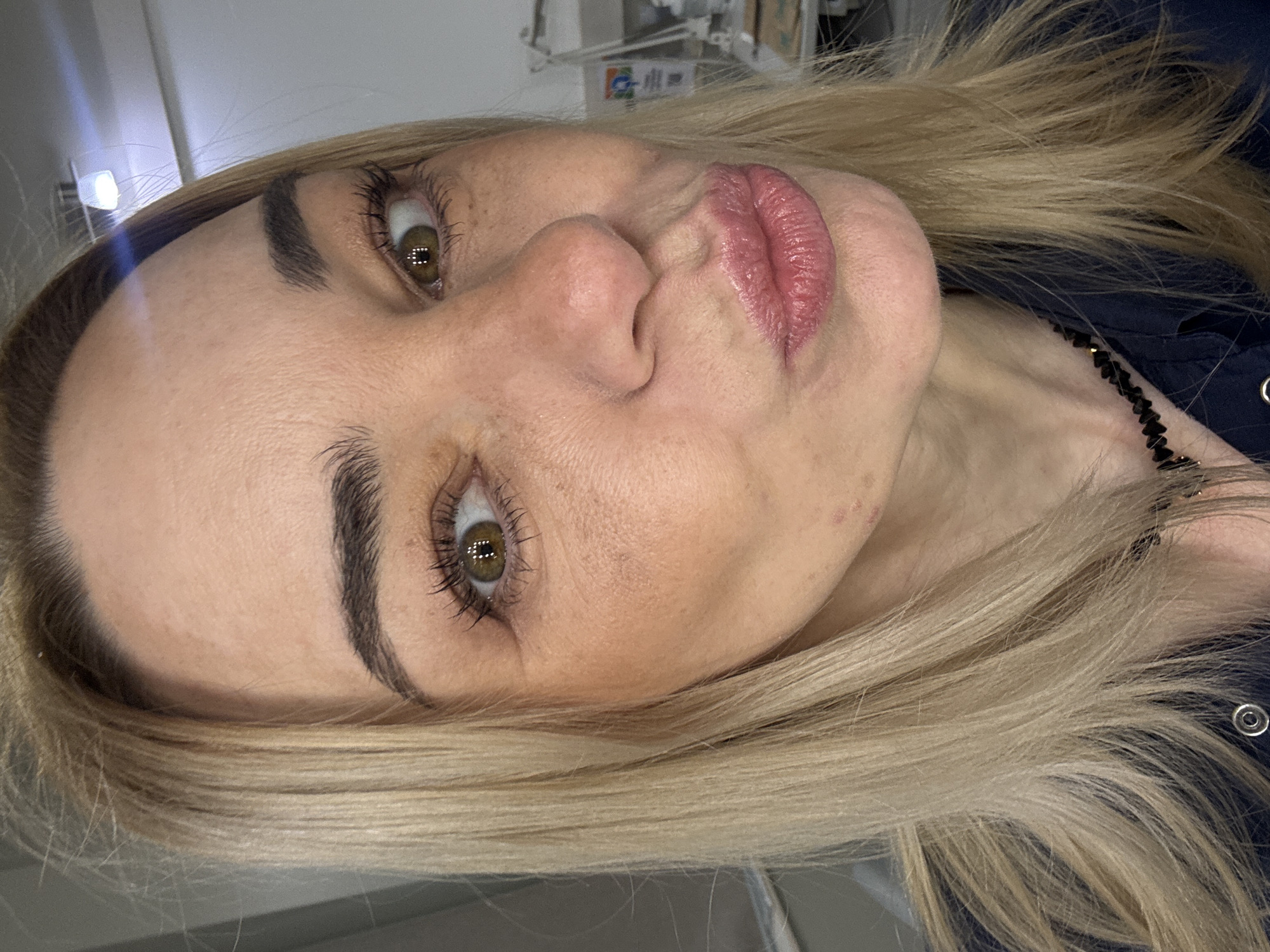 A selfie of a blonde woman with light skin, dark eyebrows and brown eyes. She is puckering her lips towards the camera. She is wearing a dark blue shirt and a black beaded necklace. The background is blurry.
