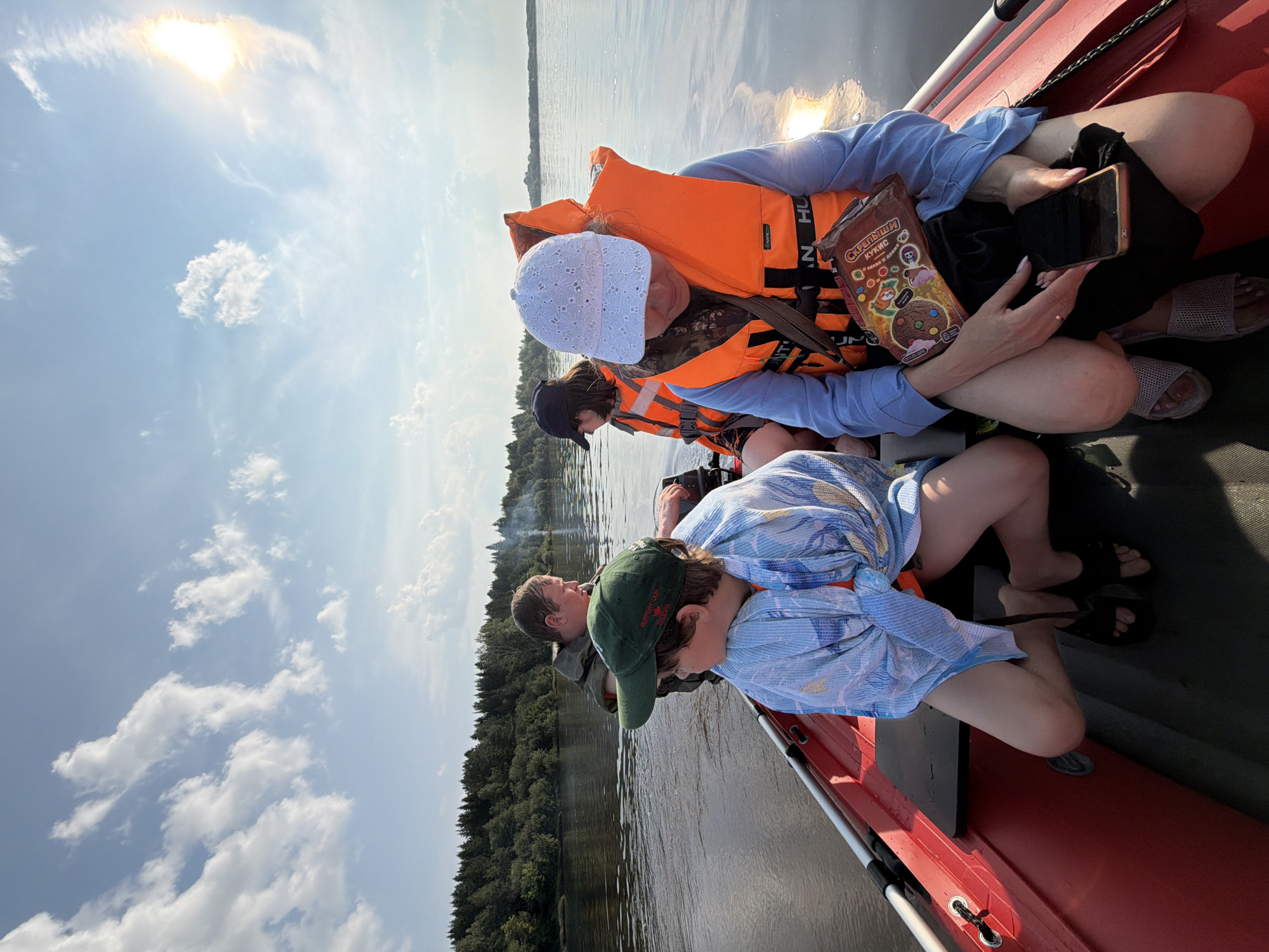 Four people are in a red motorboat on a body of water. The boat is moving, as indicated by the wake behind it. The sky is partly cloudy with the sun visible. On the left side of the boat, a person wearing a green baseball cap and a blue and white patterned garment is looking to their left. Behind them, another person in a green vest is looking forward. On the right side of the boat, a person wearing a white cap and an orange life vest is looking down at a phone and a snack bag. Next to them, another person in an orange life vest is also looking forward. The boat is passing a densely wooded shoreline. The scene appears to be in a rural area.